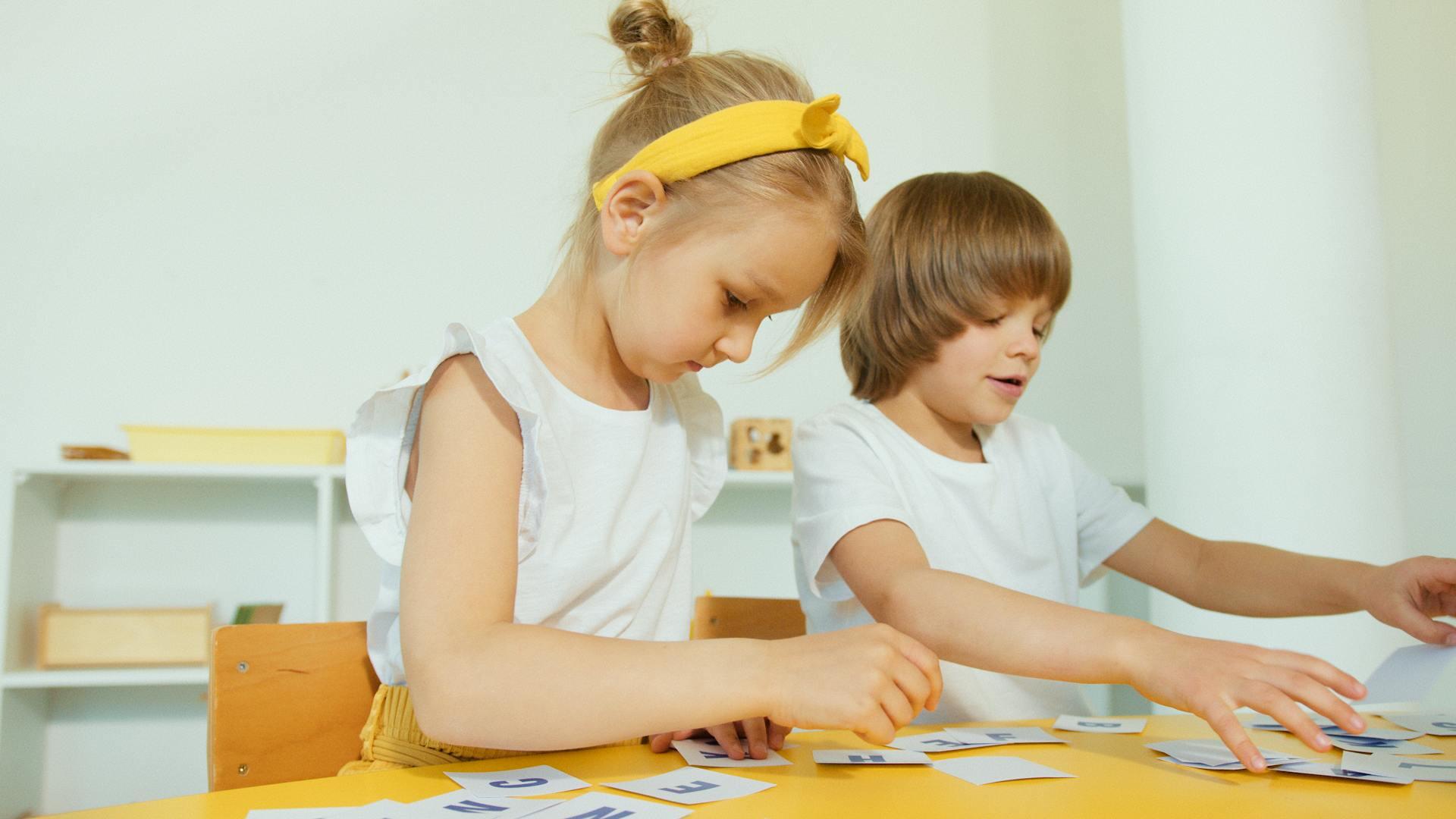 Imagen de Pexels de dos niños en un aula Montessori trabajando concentrados con tarjetas de lenguaje sobre una mesa amarilla. Foto de Artem Podrez.