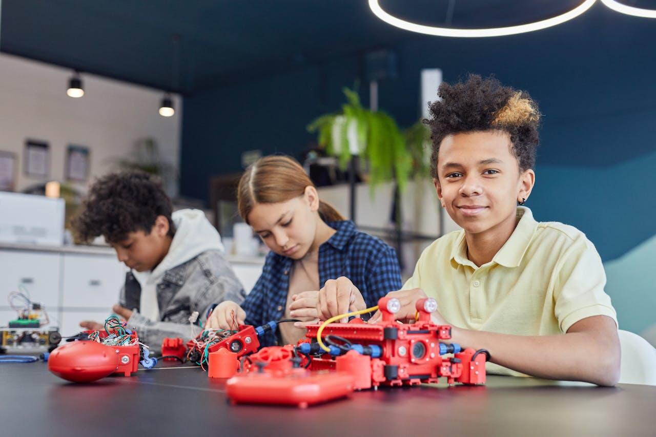 En un taller de tecnología, tres adolescentes trabajan en equipo construyendo un robot rojo; el del frente sonríe a la cámara. Fotografía de Vanessa Loring.