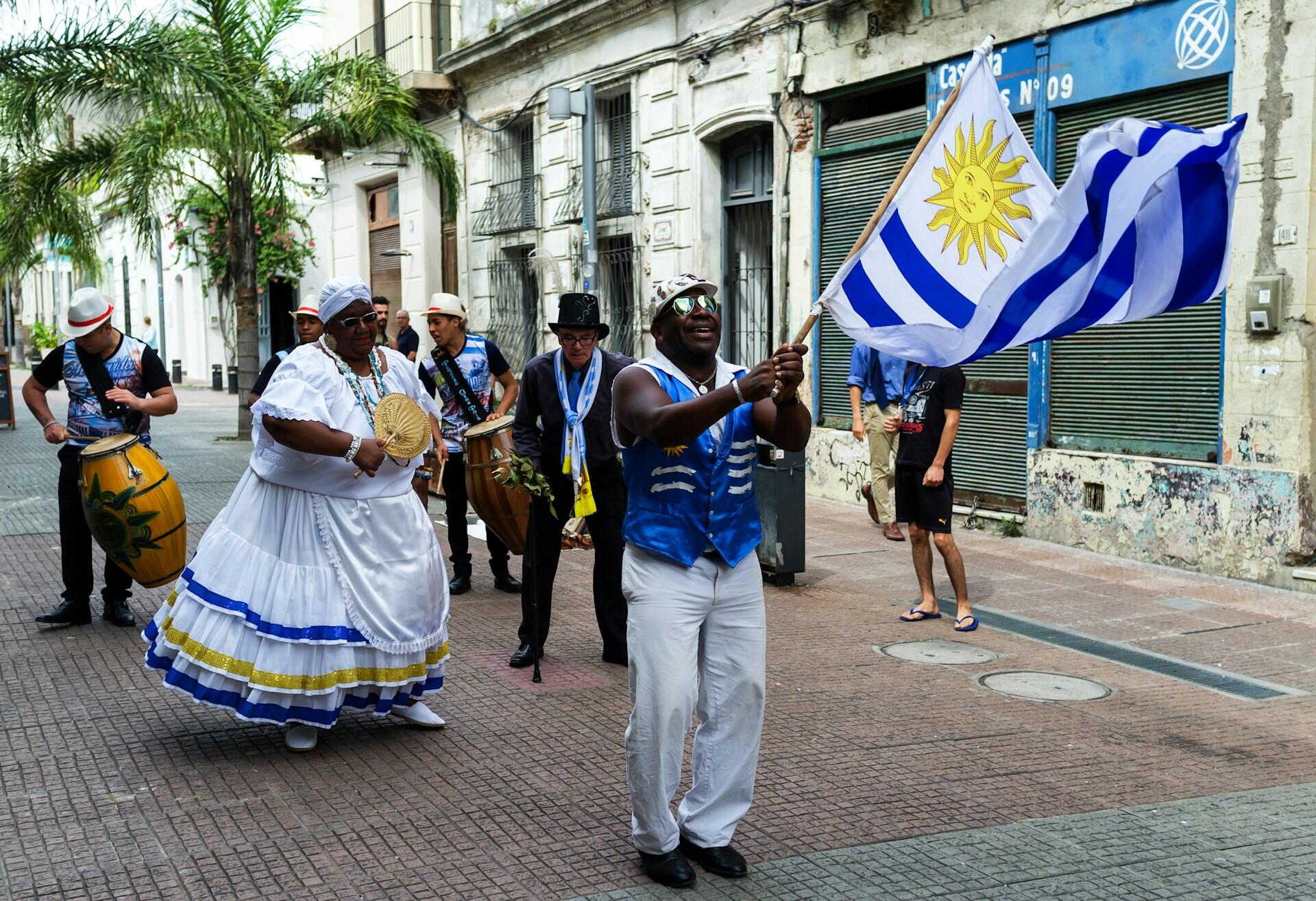 Montevideo, Uruguay. Desfile de una comparsa de candombe en la calle, con bailarines y un hombre ondeando la bandera uruguaya durante el carnaval. Fotógrafo: Nigel SB Photography.