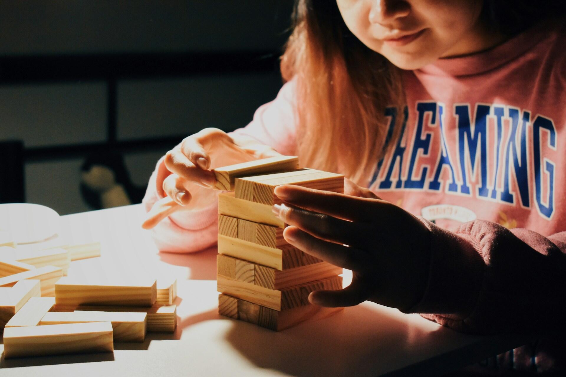 Las manos de una niña construyen con concentración una torre con bloques rectangulares de madera sobre una mesa blanca. Fotografía de Our Life in Pixels.