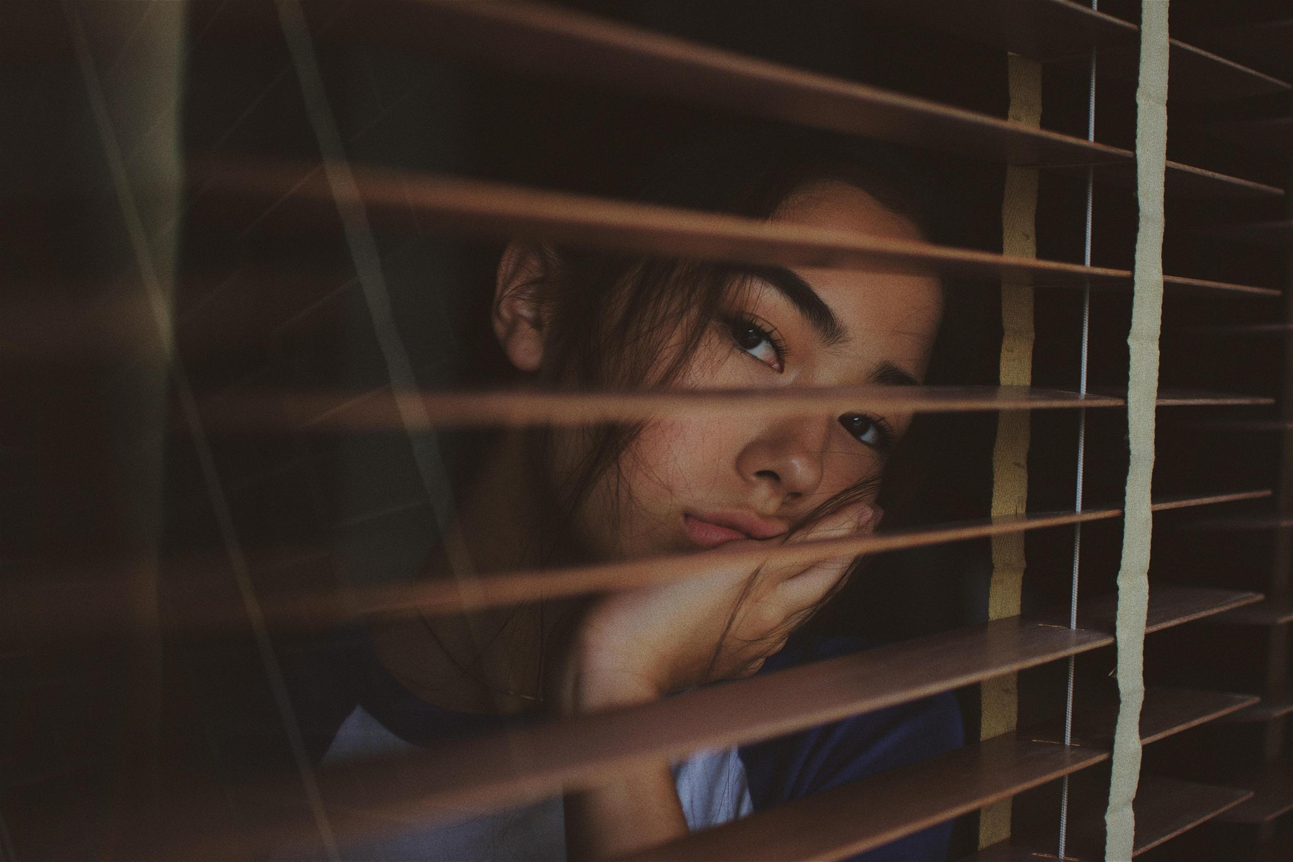 girl staring out of the window through blinds