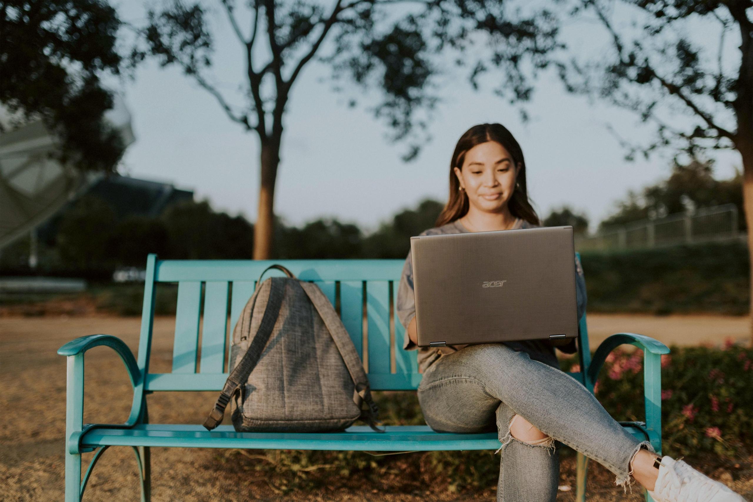 girl seated on bench next to backpack using laptop