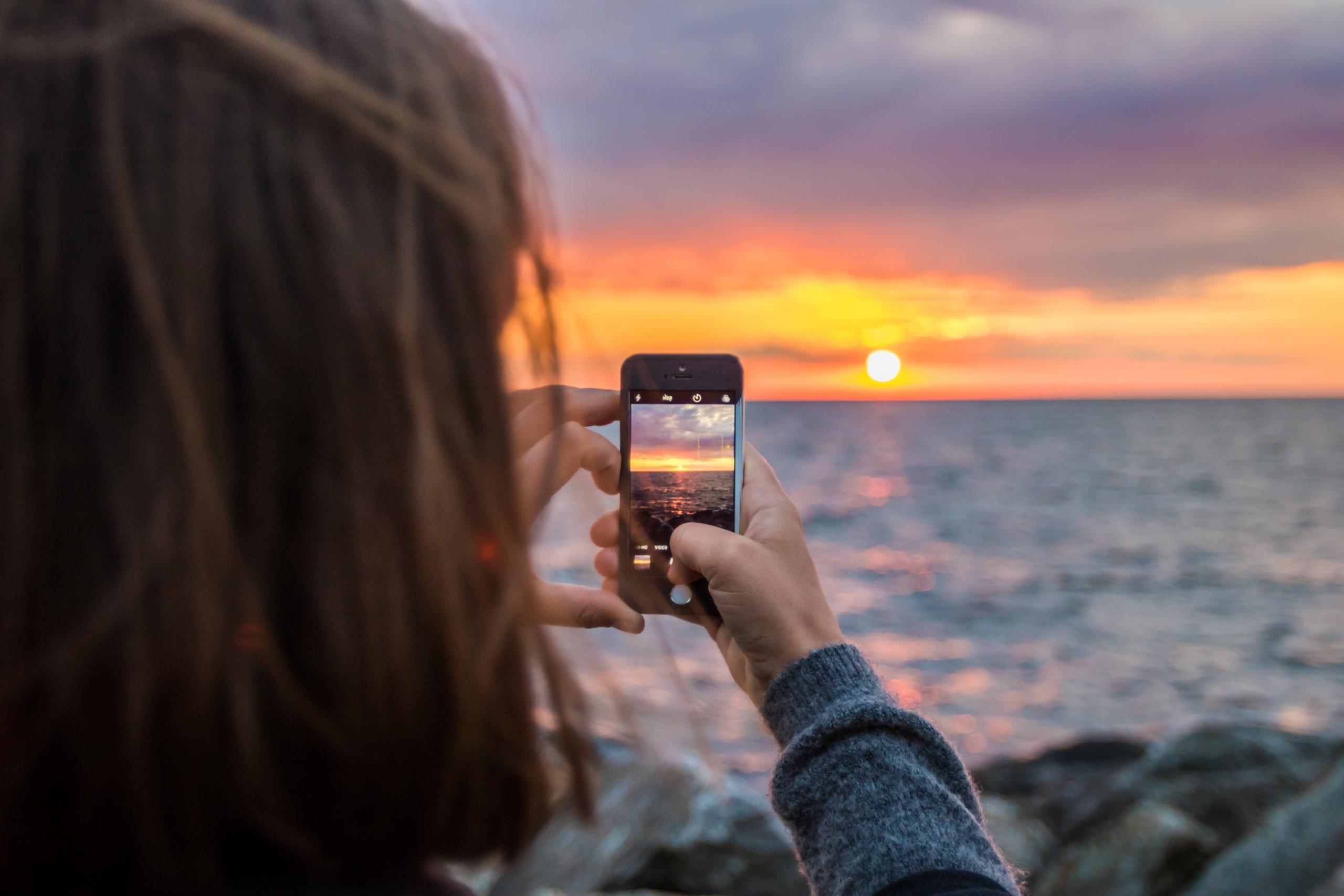 woman photographing sunset over the ocean with smartphone camera