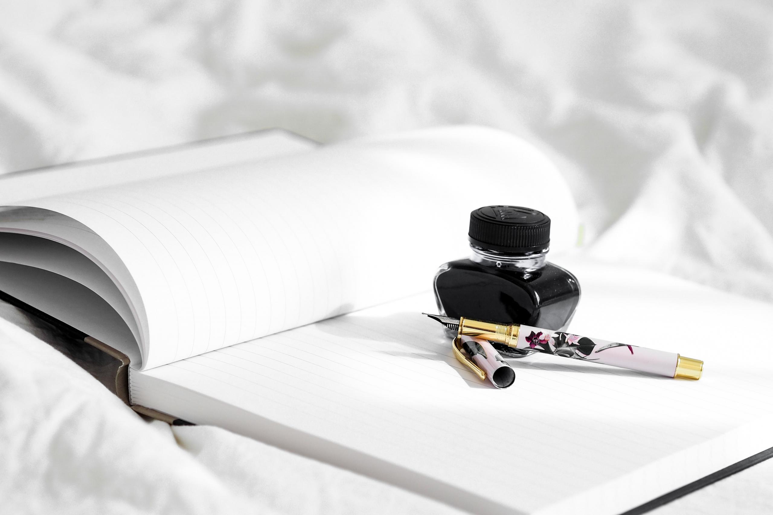 pot of ink and fountain pen placed on the open pages of an unlined notebook