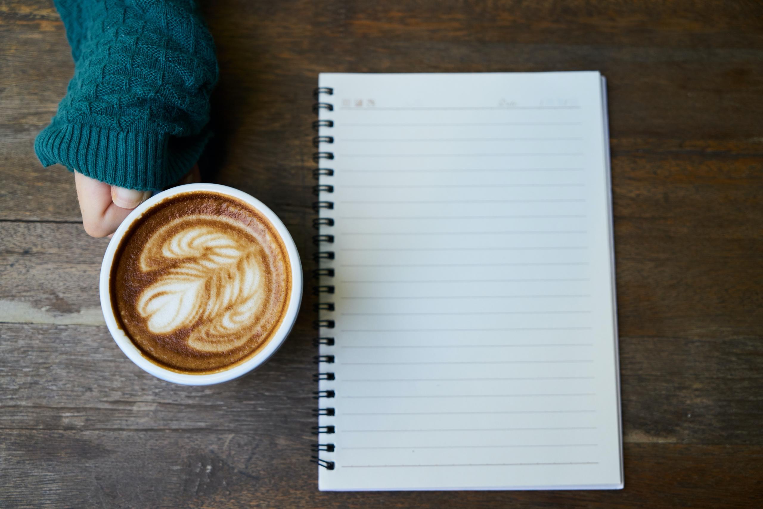 person holding coffee cup next to a blank notepad on wooden table