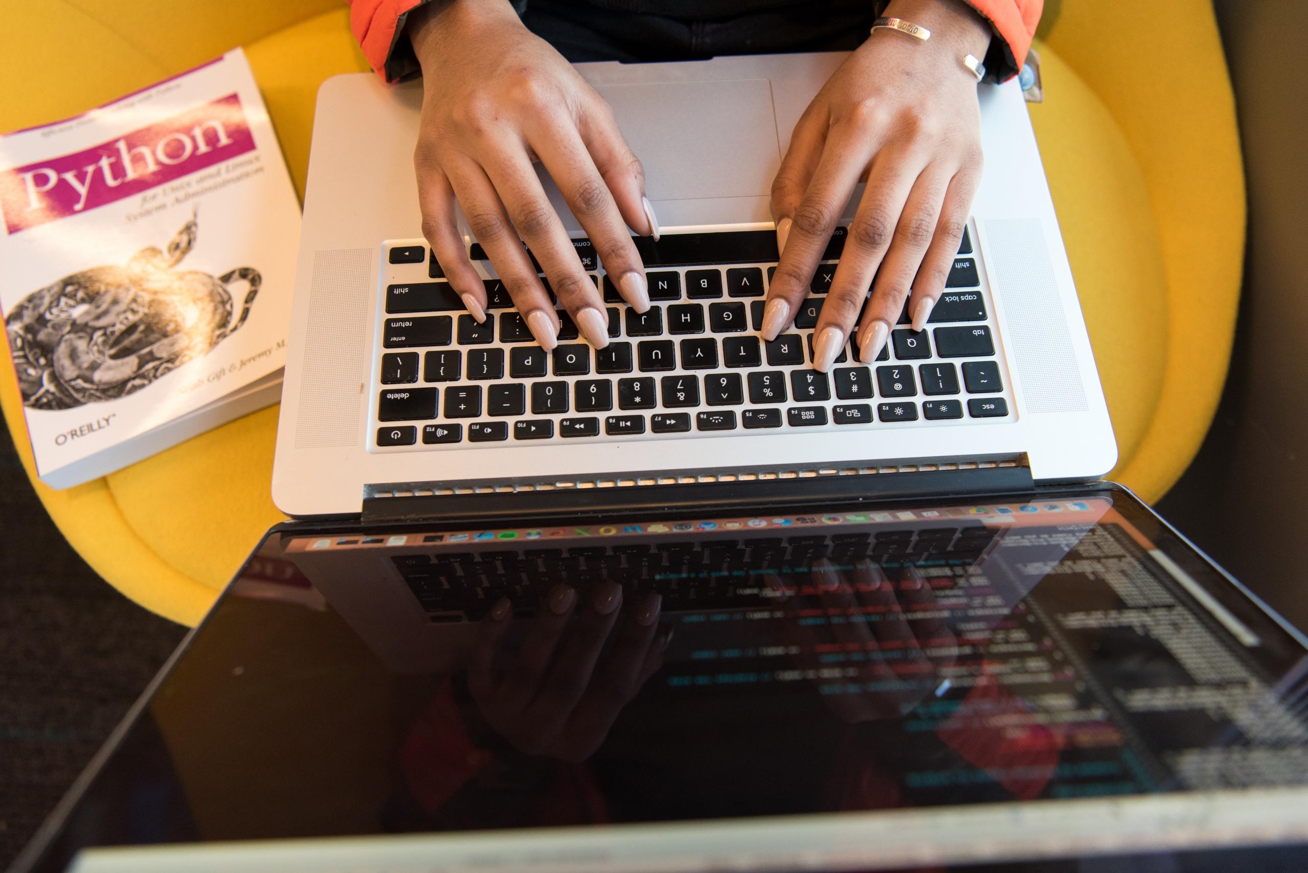 person coding Python on laptop with manual placed on chair