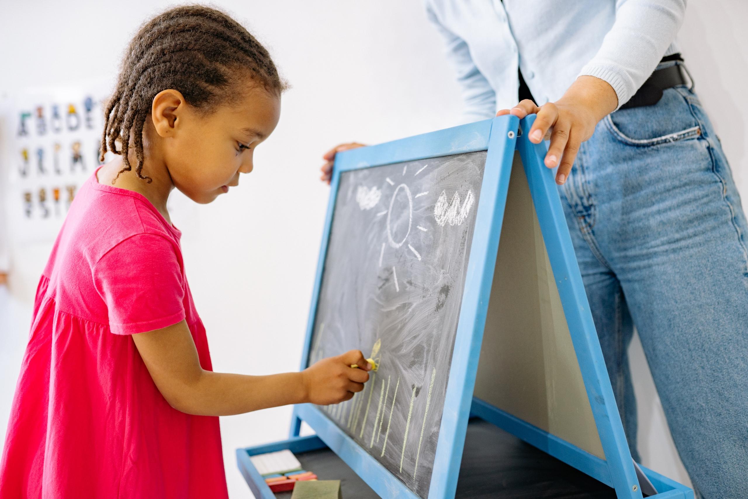 pre-schooler drawing on small chalkboard while teacher looks on