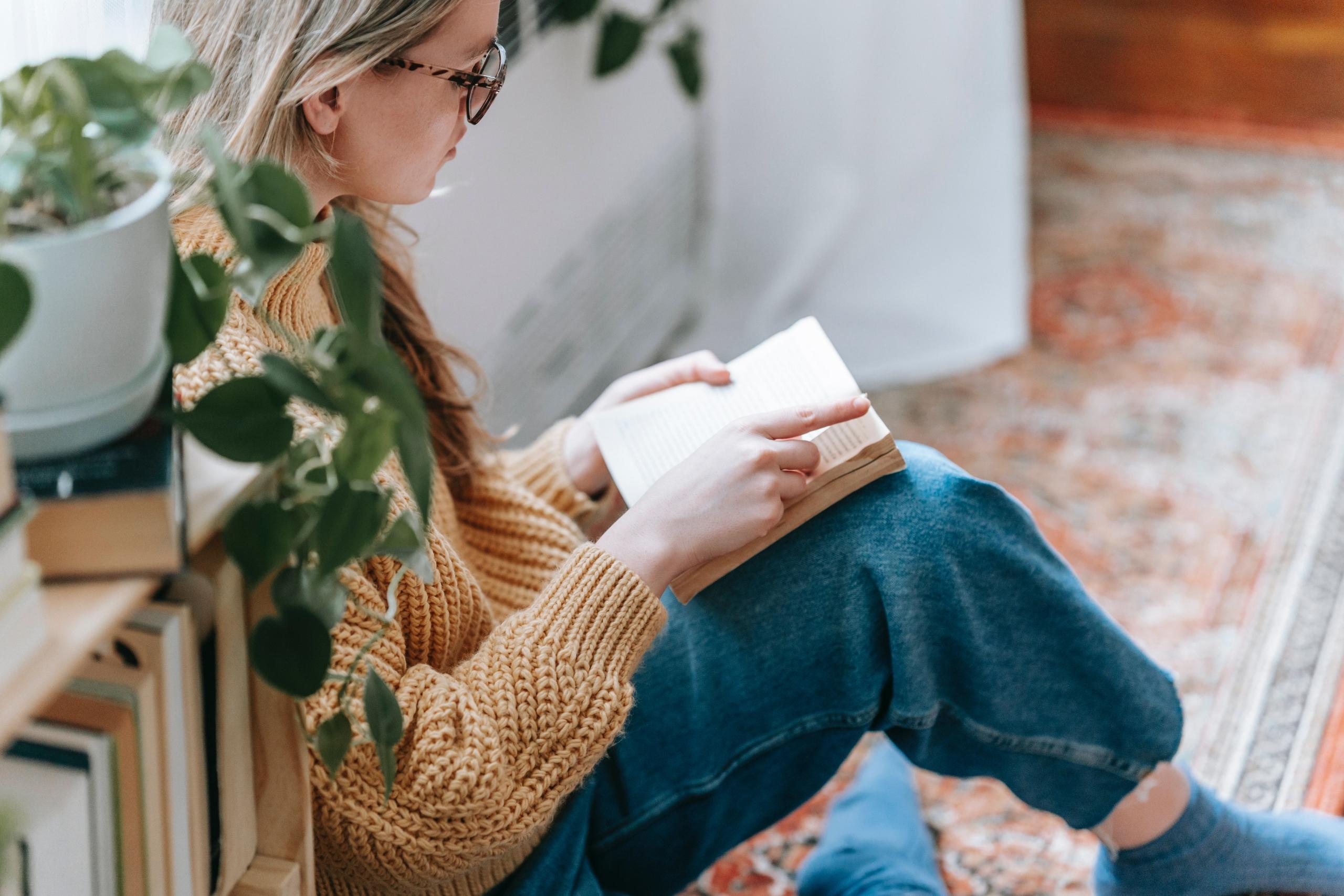 woman wearing mustard yellow pullover sitting on the floor and reading a book
