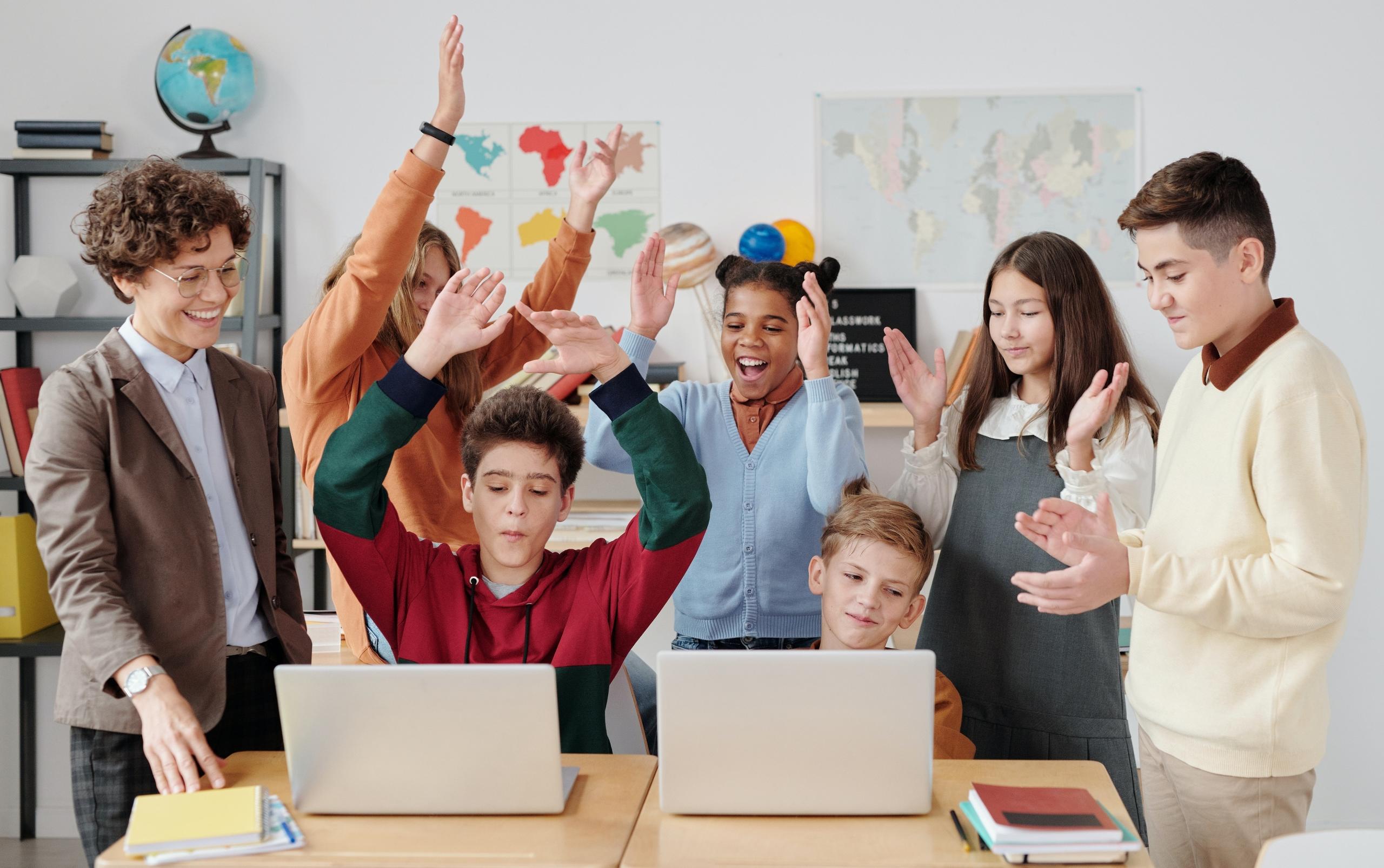 young students huddled behind two laptops in classroom celebrating