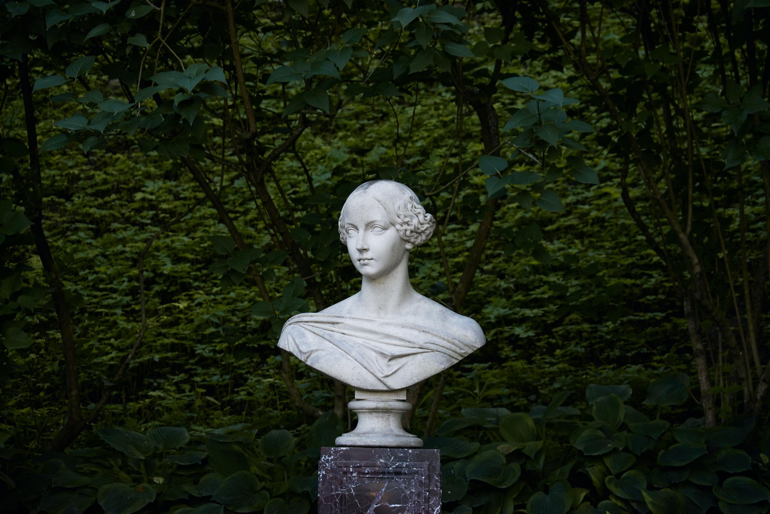 bust of a woman on a plinth in front of trees and a green hedge