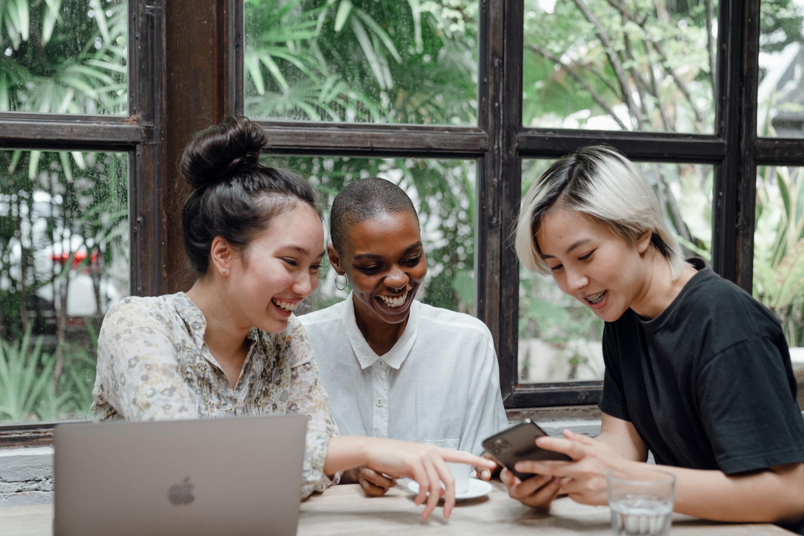 multi cultural group of women around table laughing using digital devices