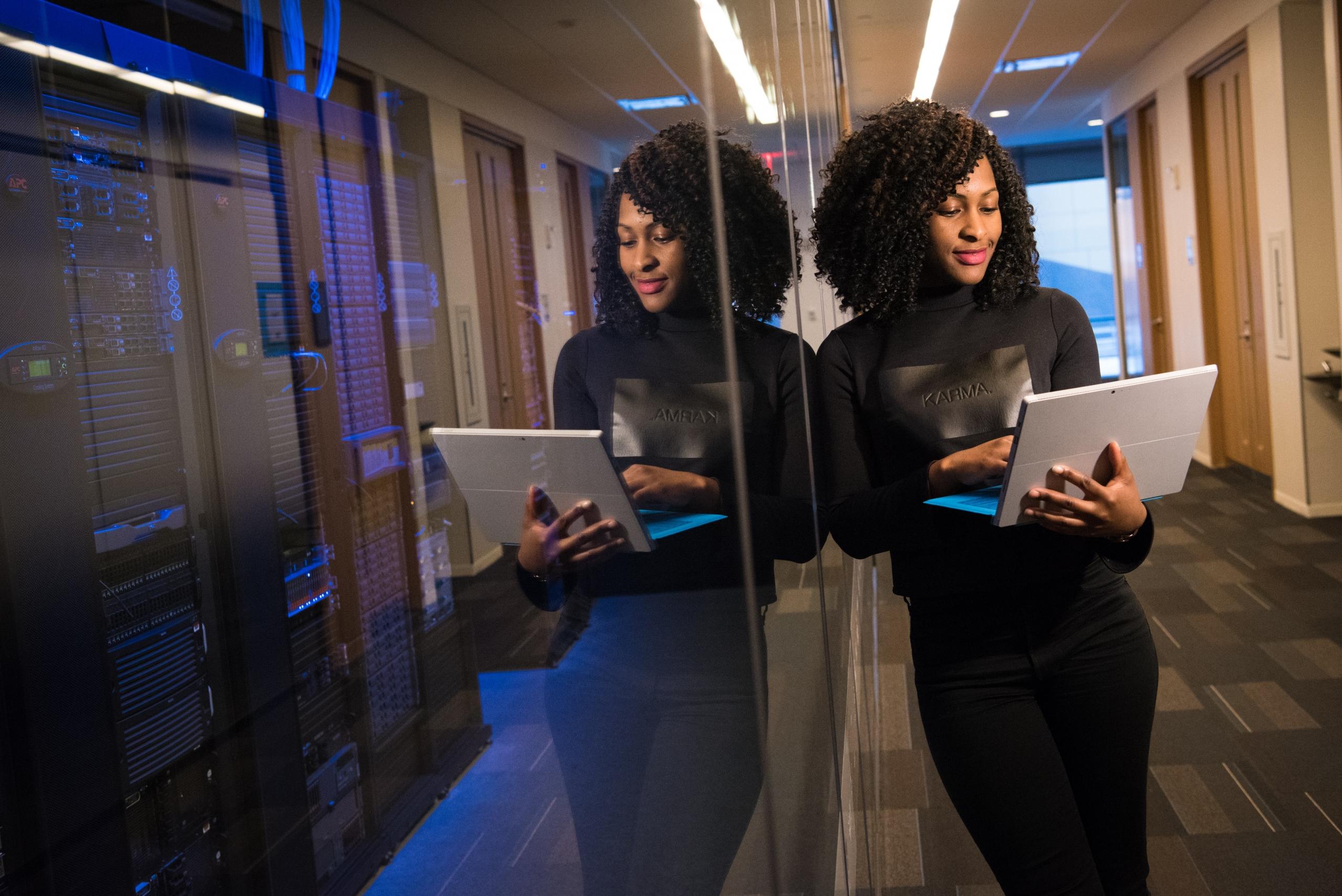 businesswoman leaning against glass office wall while typing on laptop