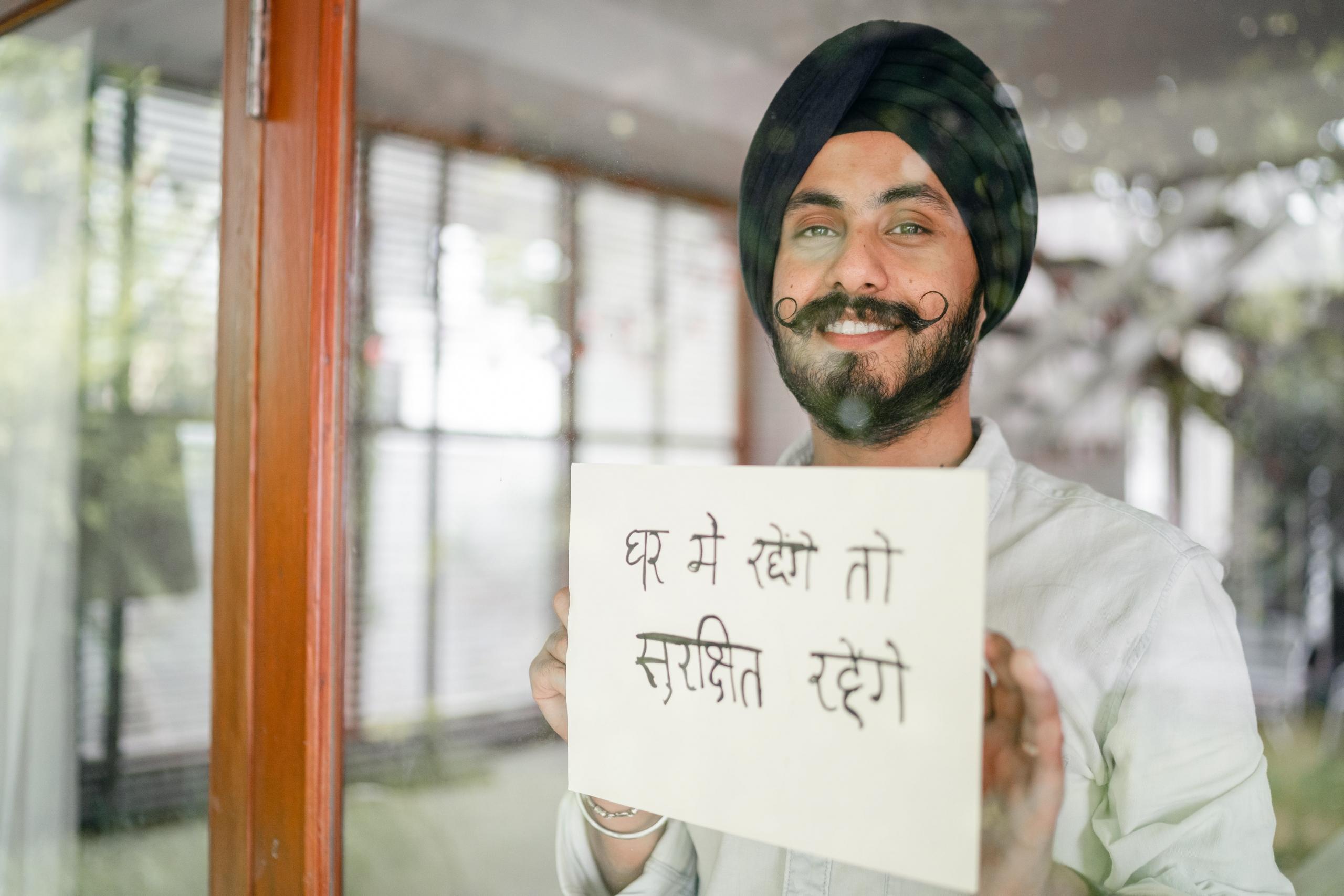 smiling man wearing turban and holding sign with hindi text up to window