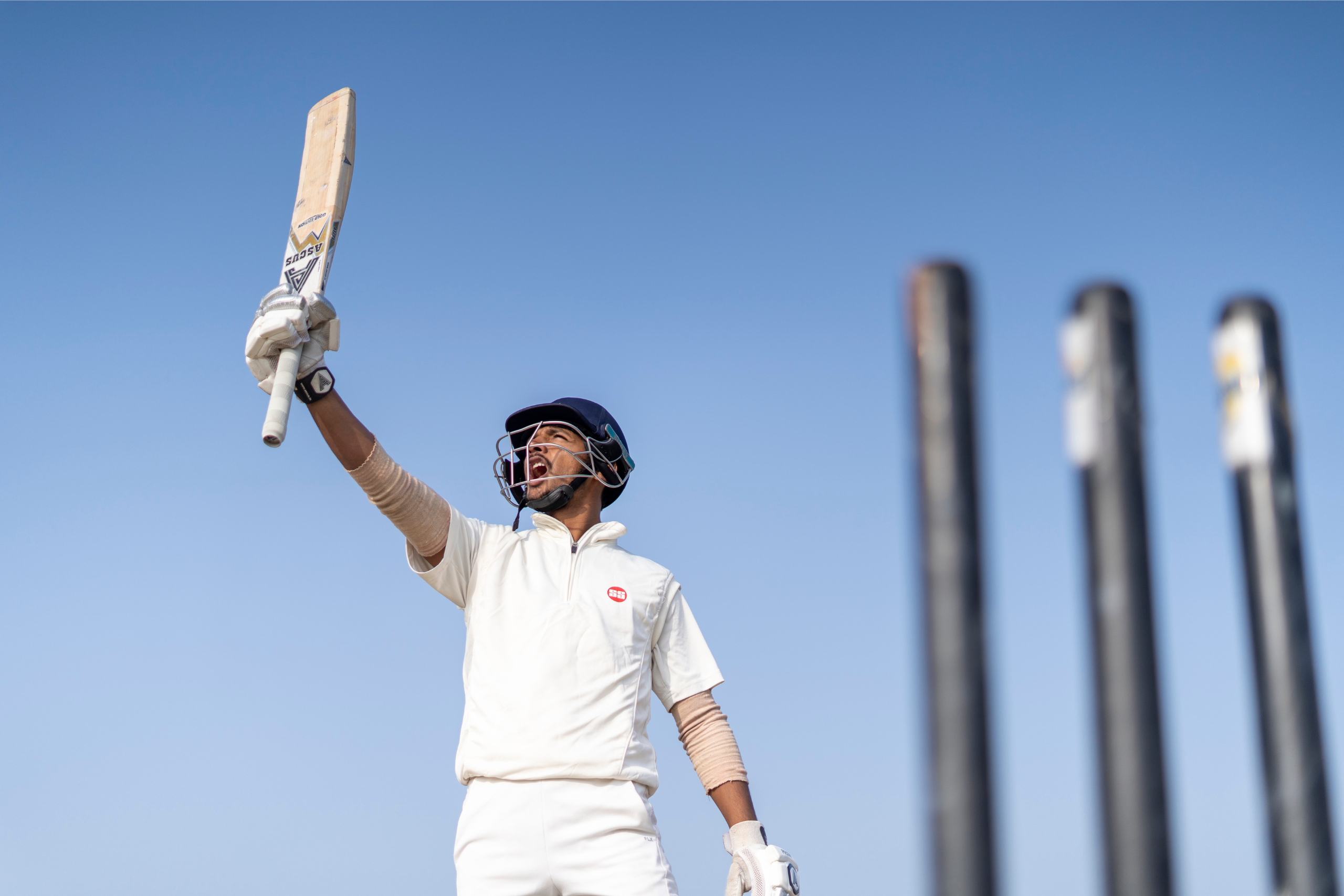 cricket player standing behind wickets raising his cricket bat in the air under blue skies