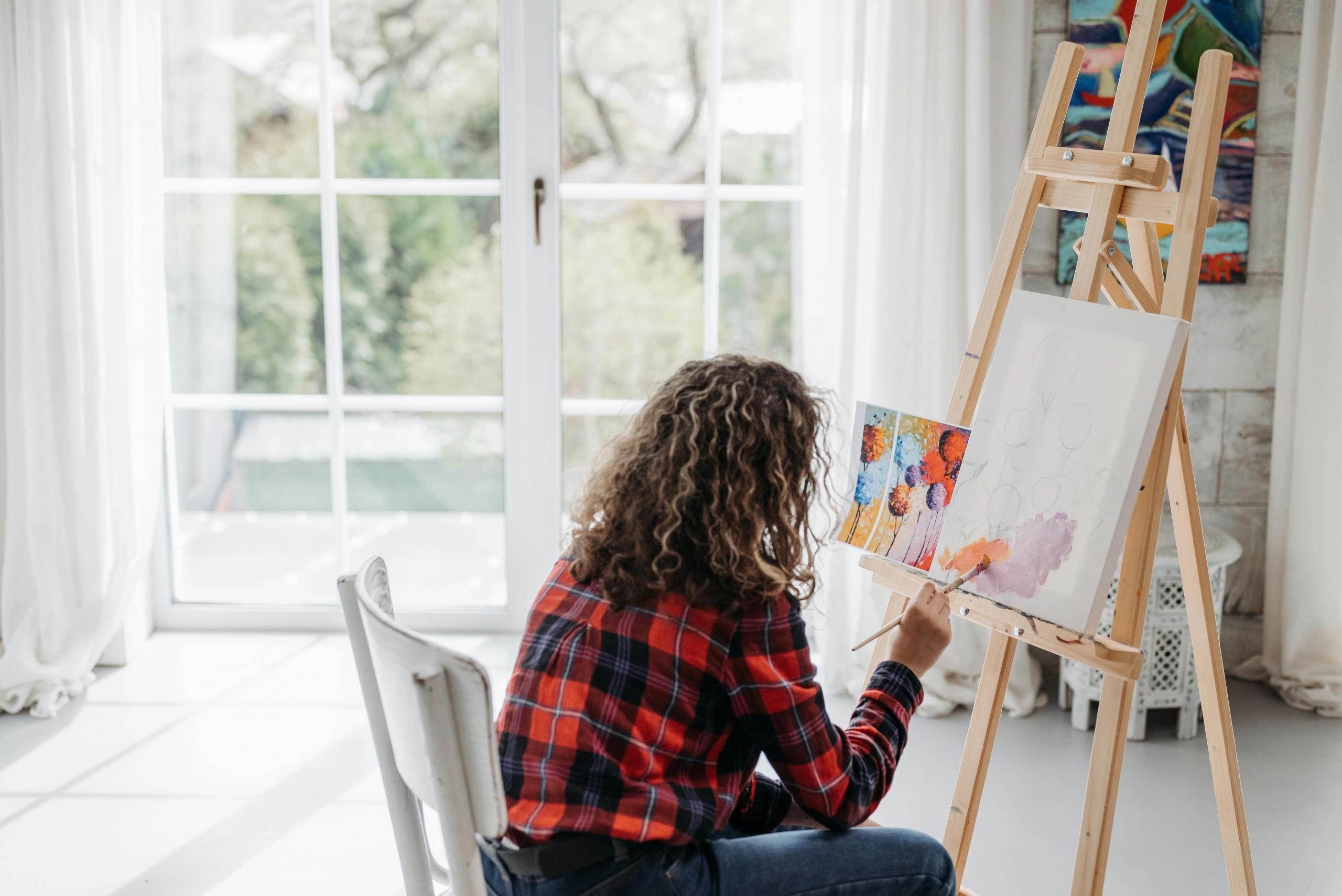 artist sitting on a chair near a window painting on a canvas balanced on easel