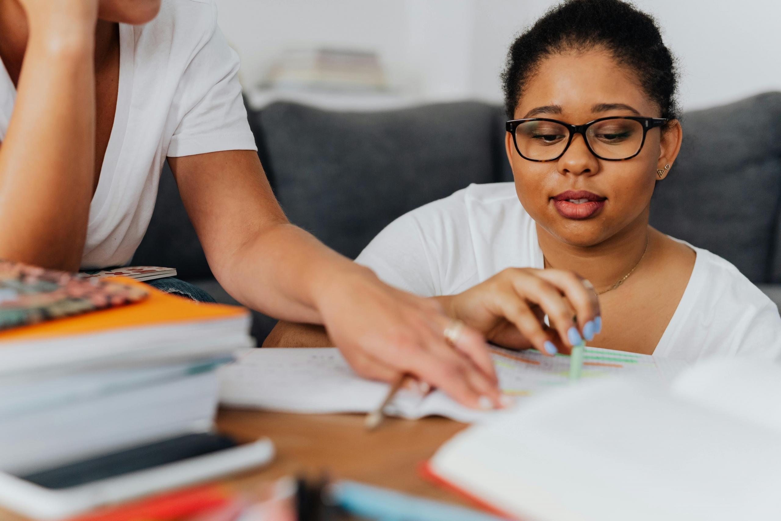 student sitting on floor doing maths exercises in notebook on coffee table while tutor guides her