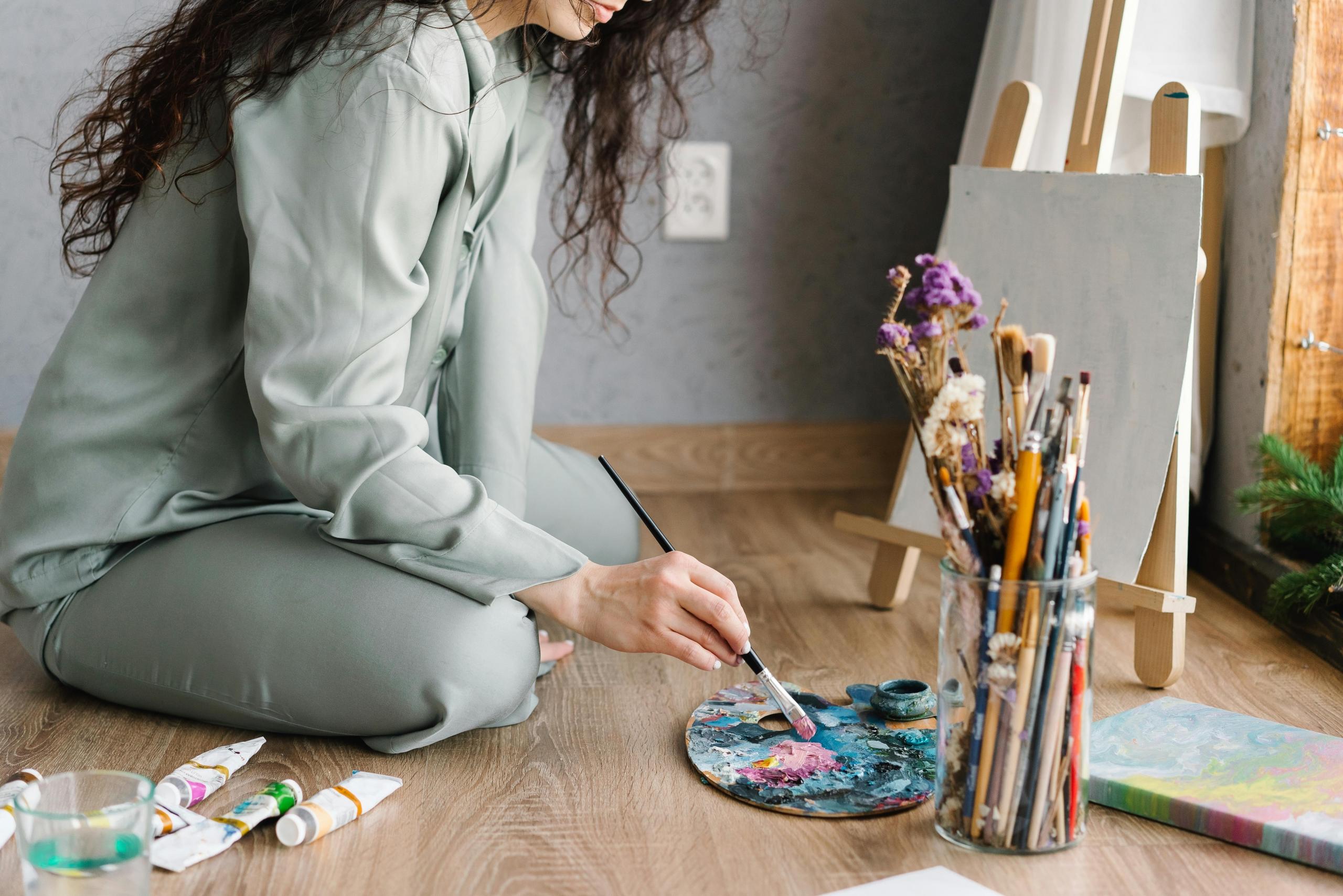 woman sitting on the floor in front of blank canvas on easel brushing paintbrush across palette of paint