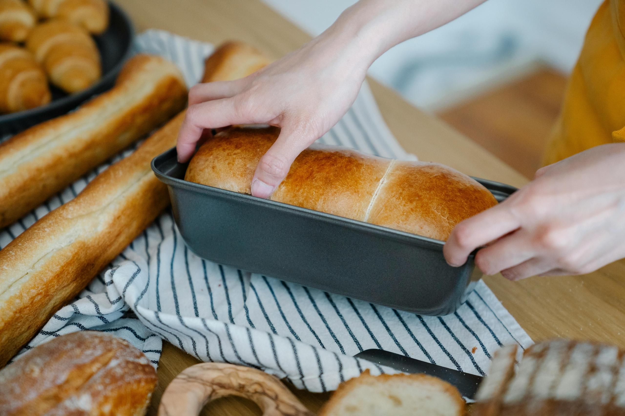 baker taking a loaf of bread out of baking tin, with freshly baked croissants and baguettes in background