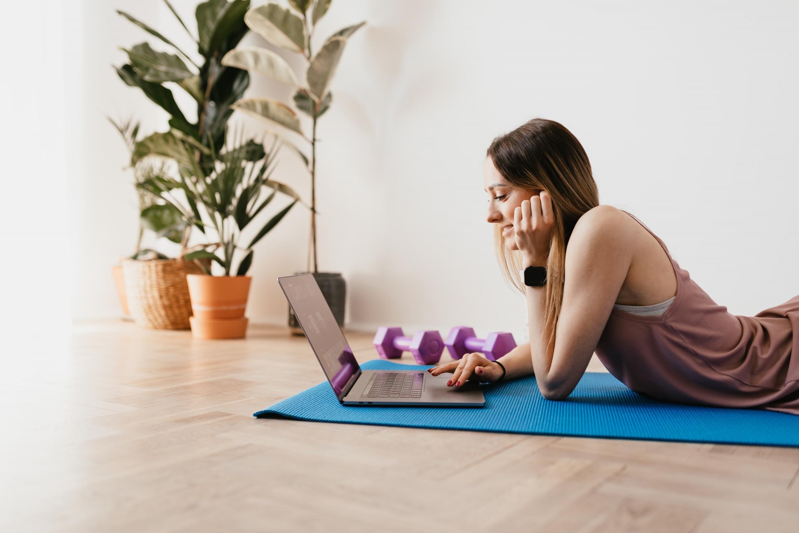 woman lying on exercise mat next to dumbbells, logging into online fitness class on laptop