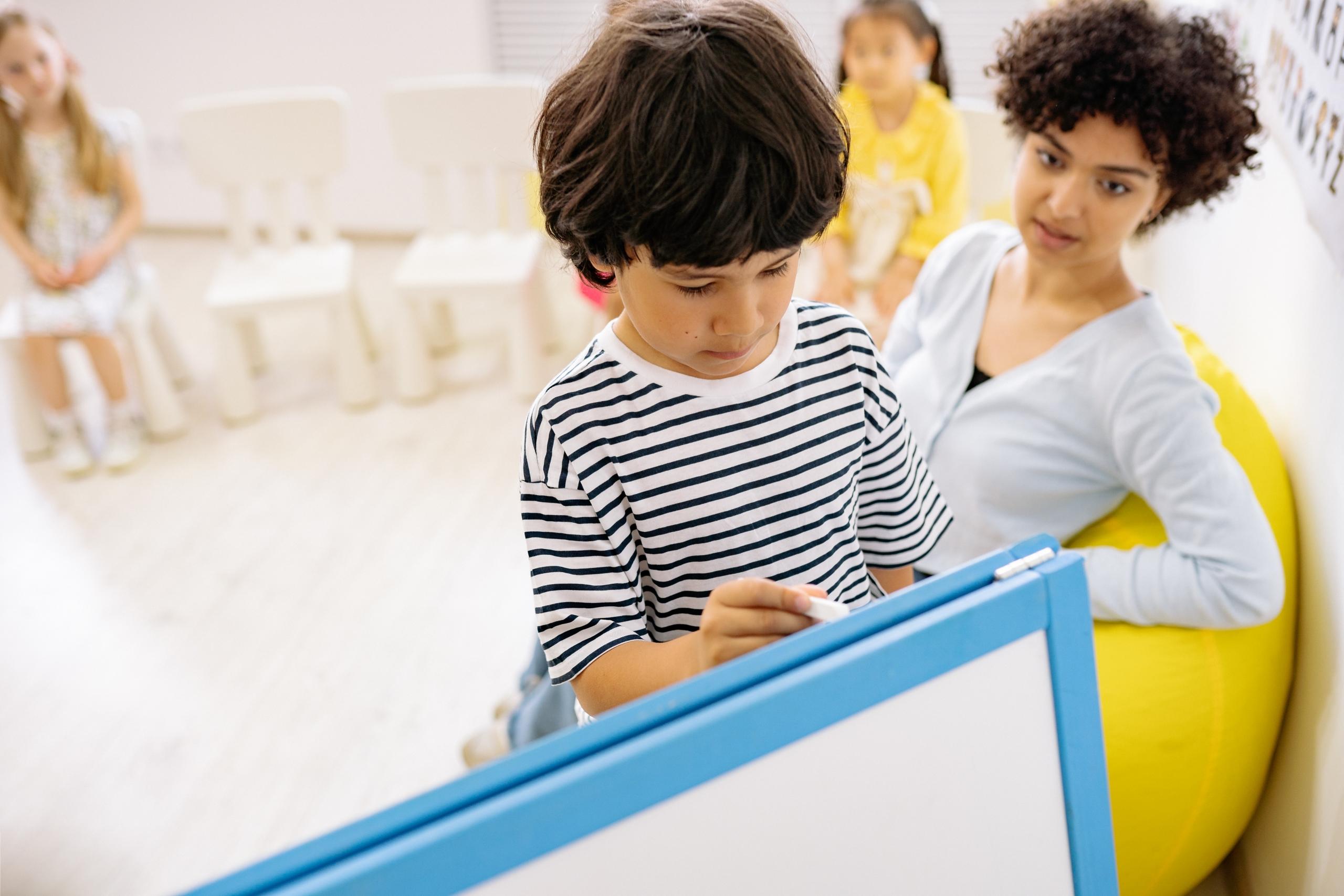 boy writing on small whiteboard while teacher supervises