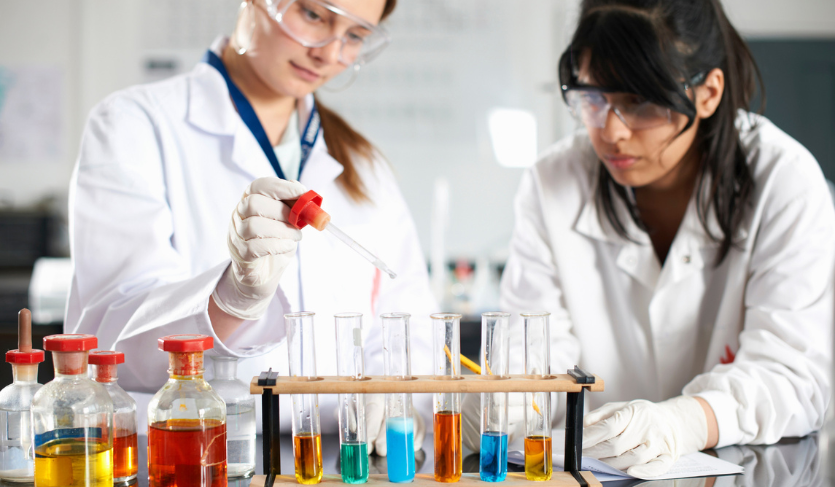 two high school students wearing lab coats and protective goggles perform chemical experiment in class