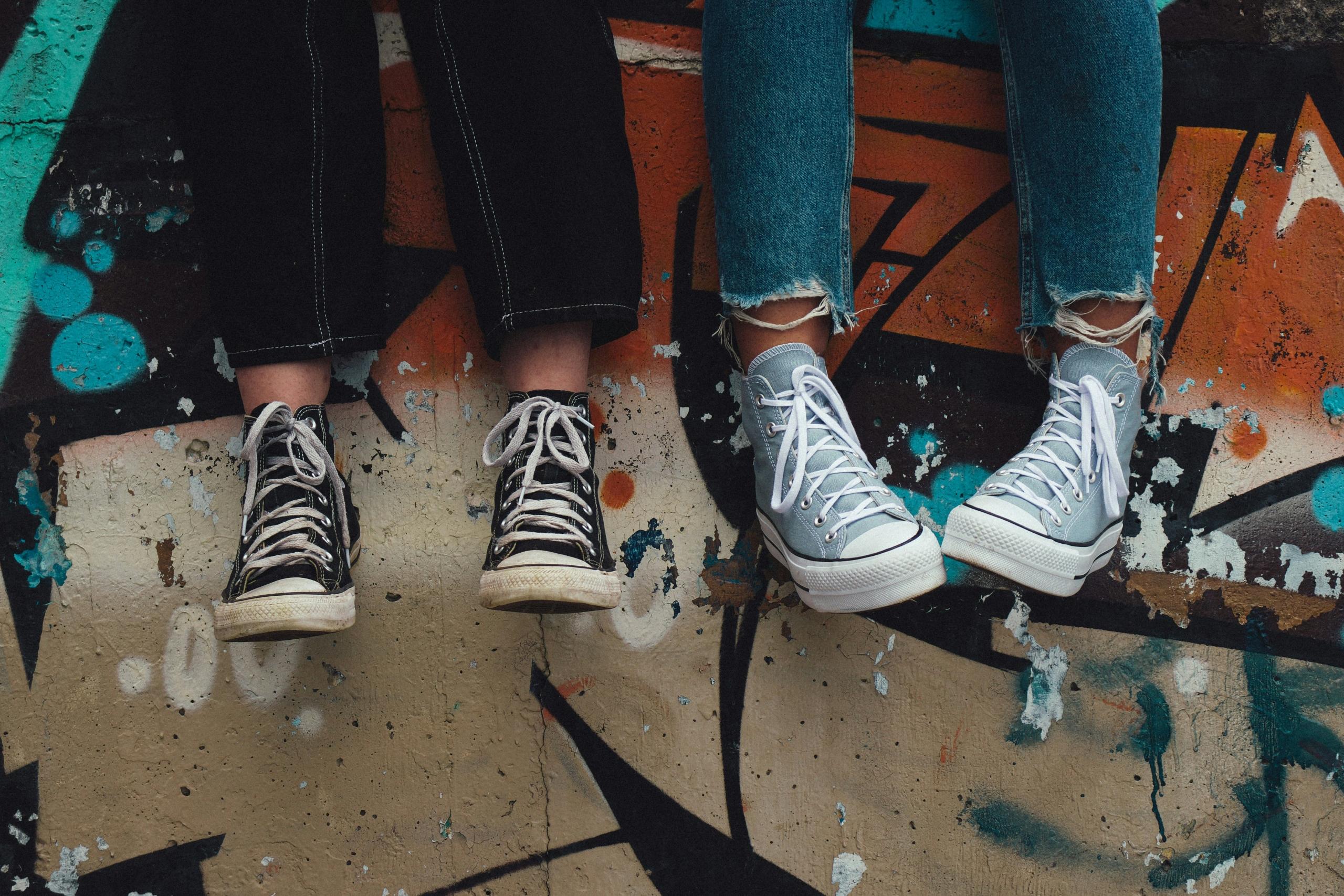 close up shot of feet of two teens wearing high tops seated on a graffiti covered wall