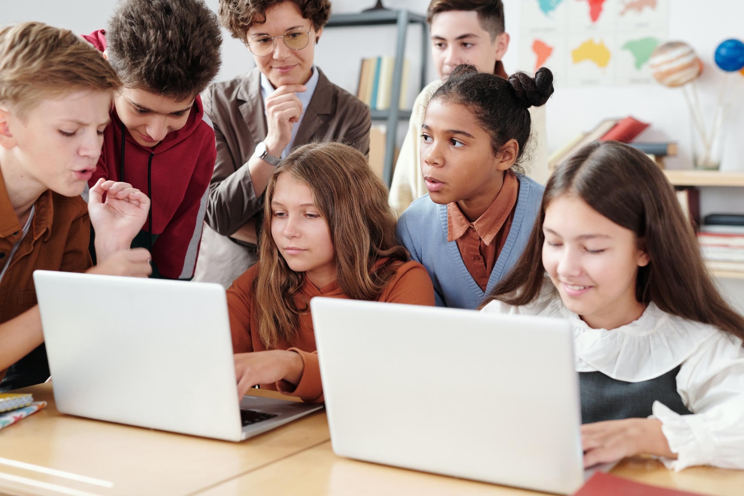 group of school learners gathered around a desk watching two girls using laptops as teacher looks on from behind
