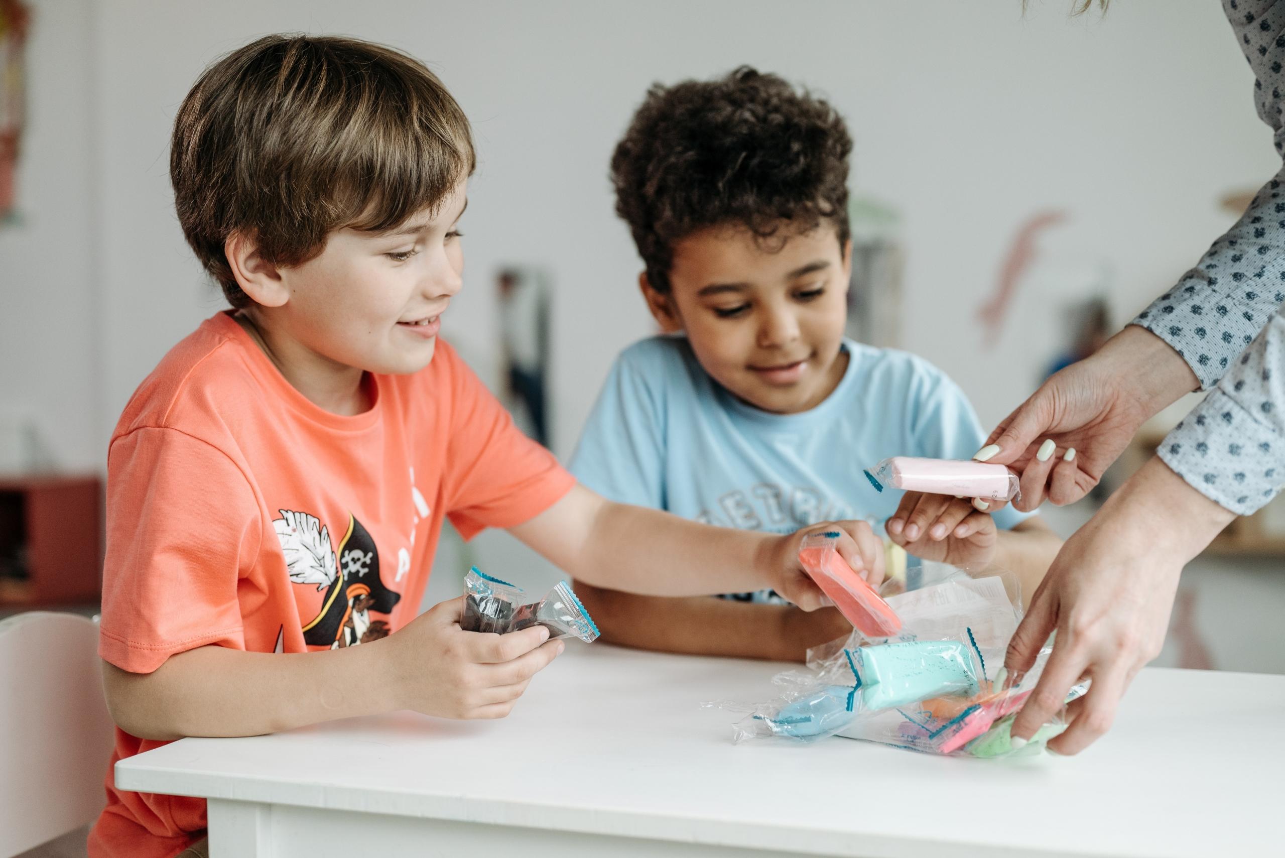 two young boys sitting at school table taking colourful packets from teacher