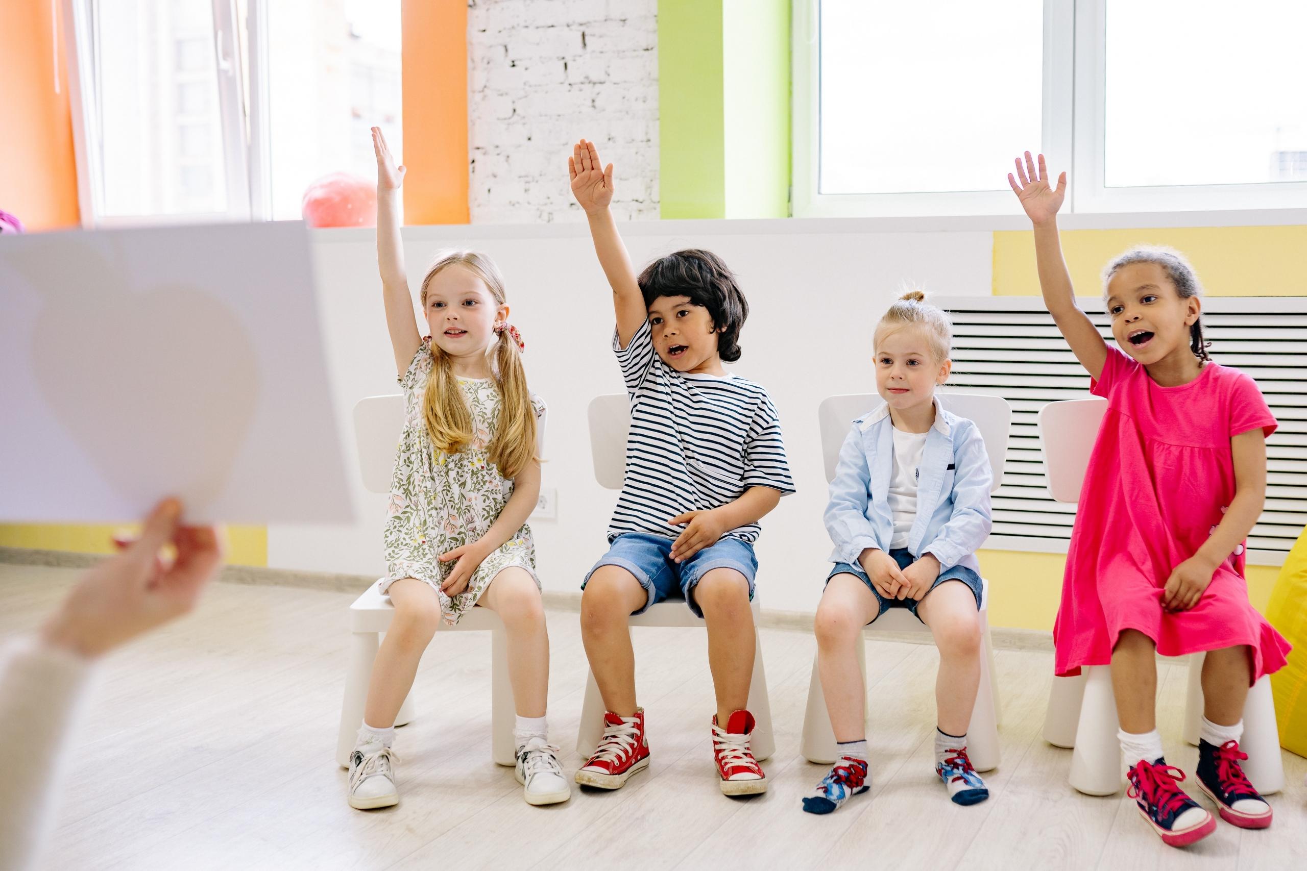 preschool children sitting side by side on chairs and raising their hands as teacher shows them flashcards