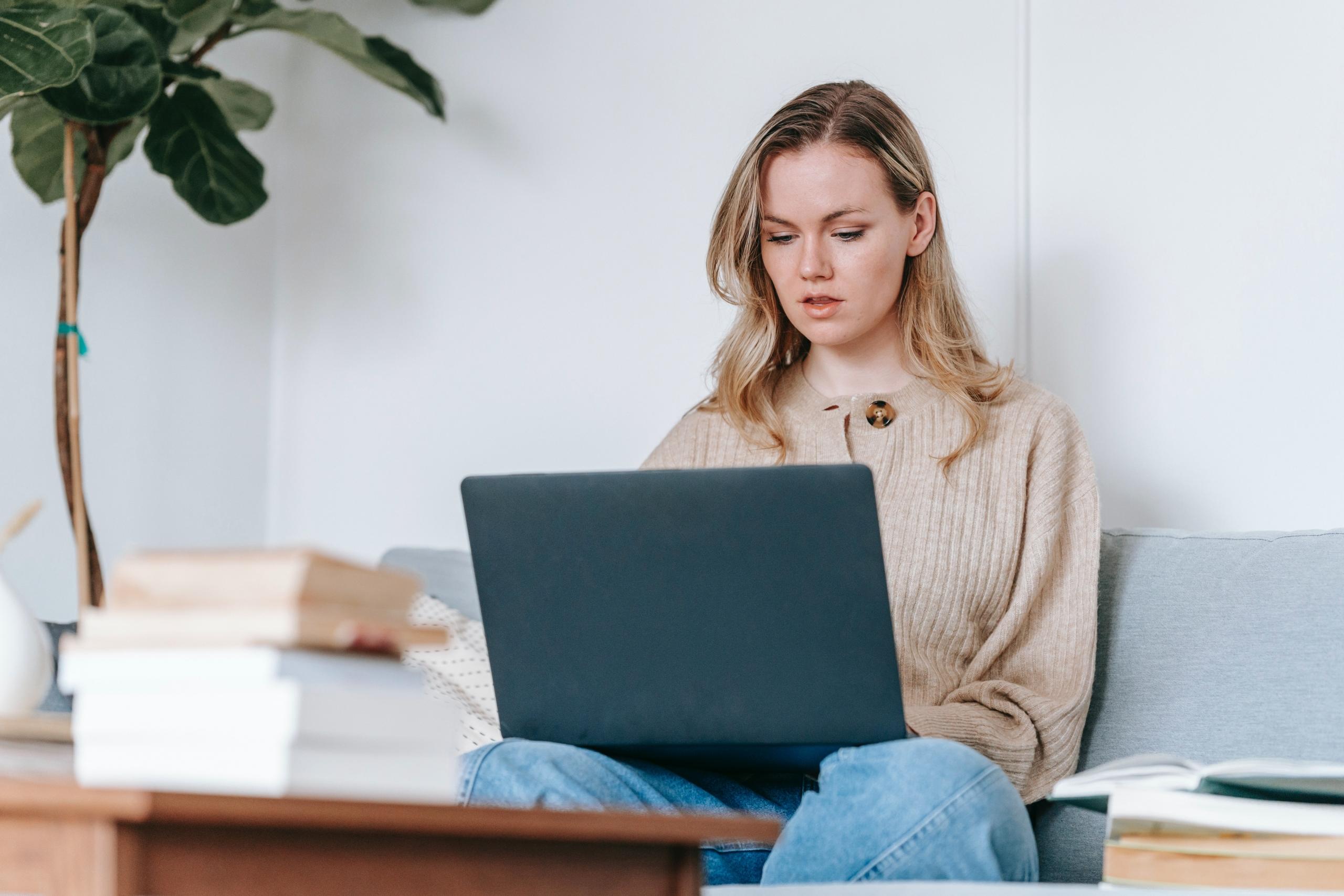 woman sitting on couch using laptop with books piled next to her and on the coffee table in front of her