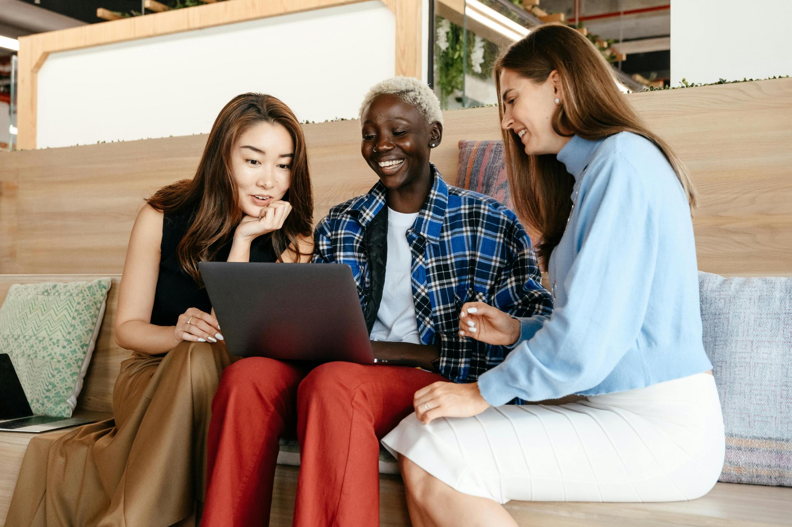 three smiling colleagues sitting side-by-side on a bench all looking at the same computer screen
