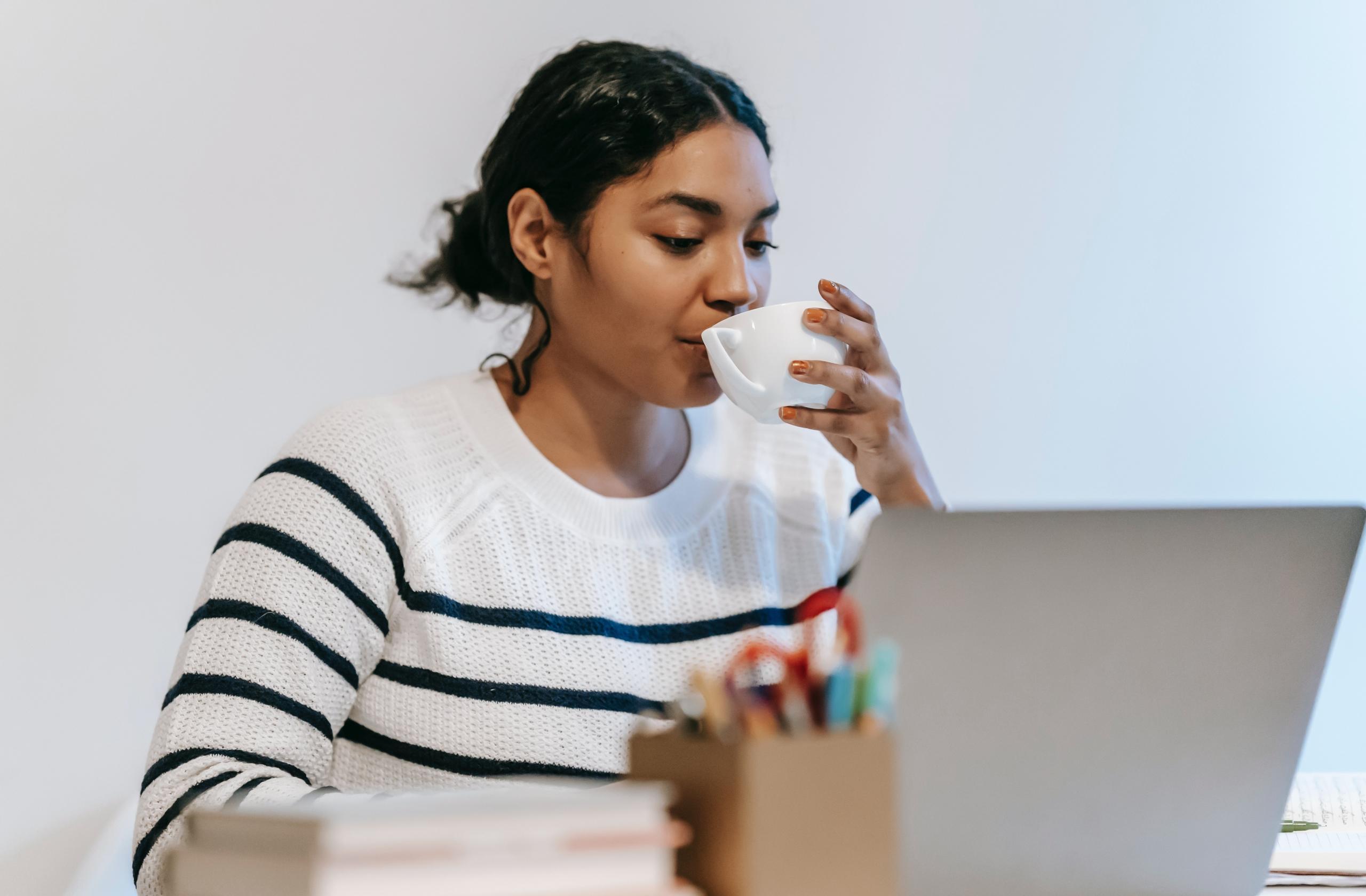 young woman sipping tea and doing work at desk on laptop with a pile of books placed alongside