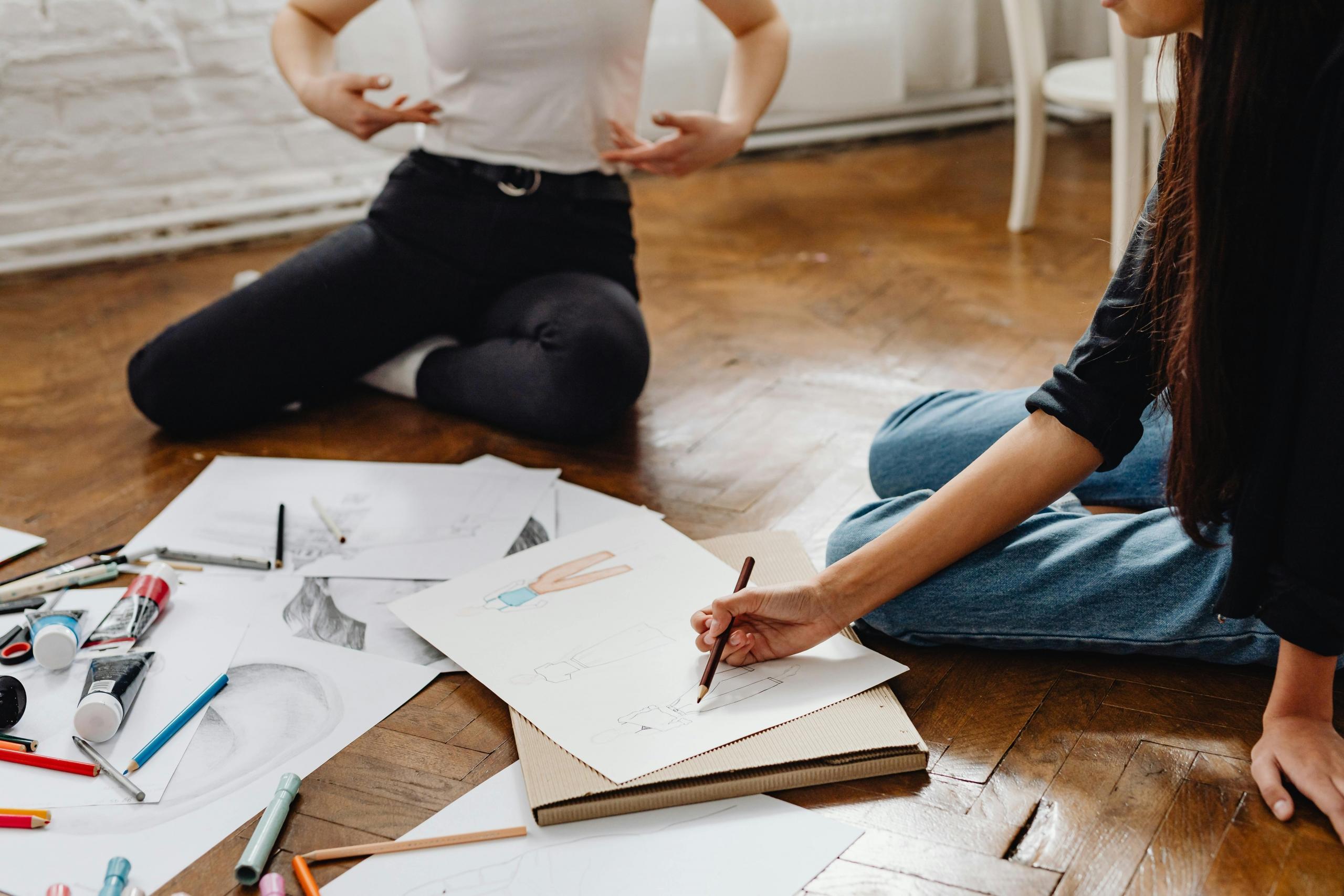 fashion design teacher showing student how to sketch sitting on wooden floor