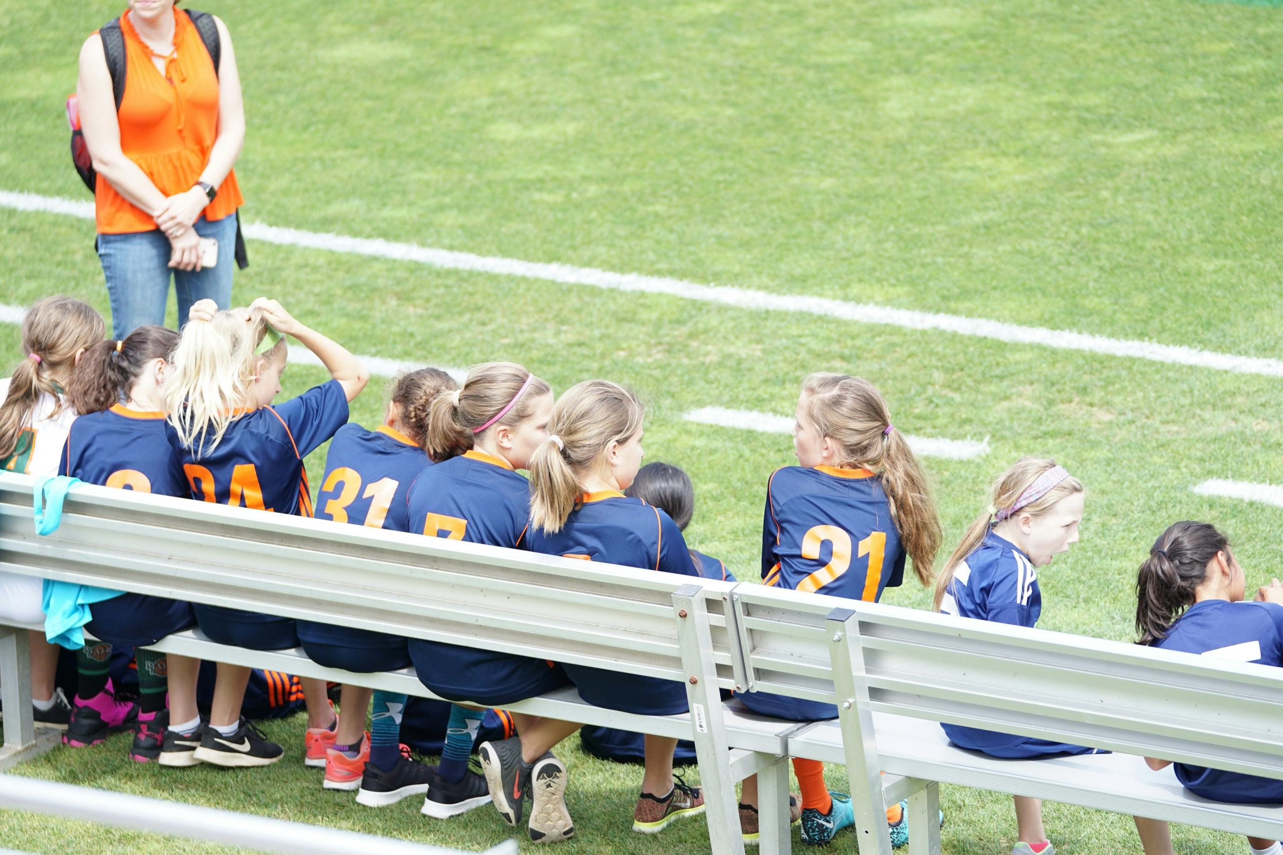 girls in soccer uniform sitting on bench next to soccer field