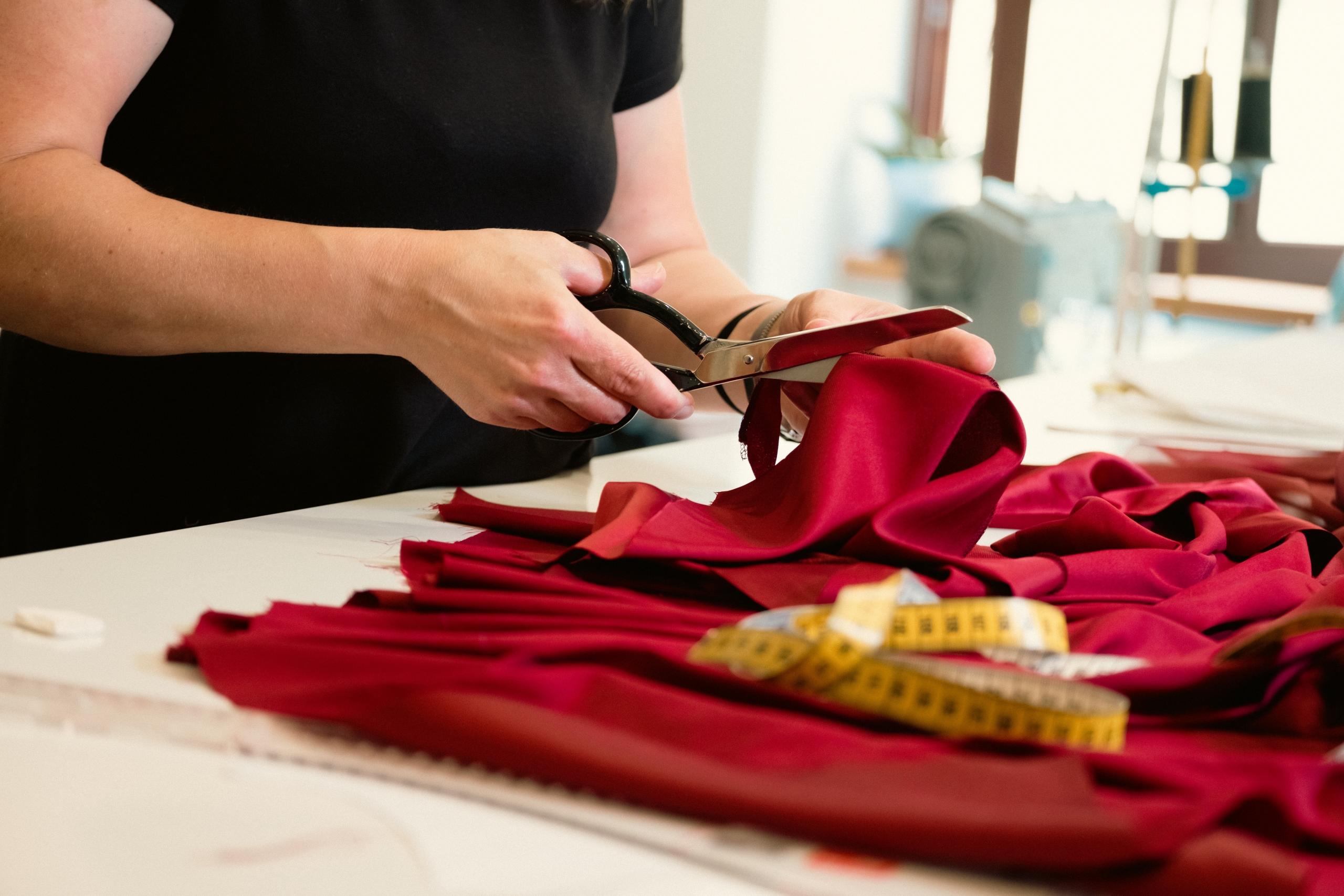 seamstress using tape measure and dressmaking scissors to cut red stain fabric