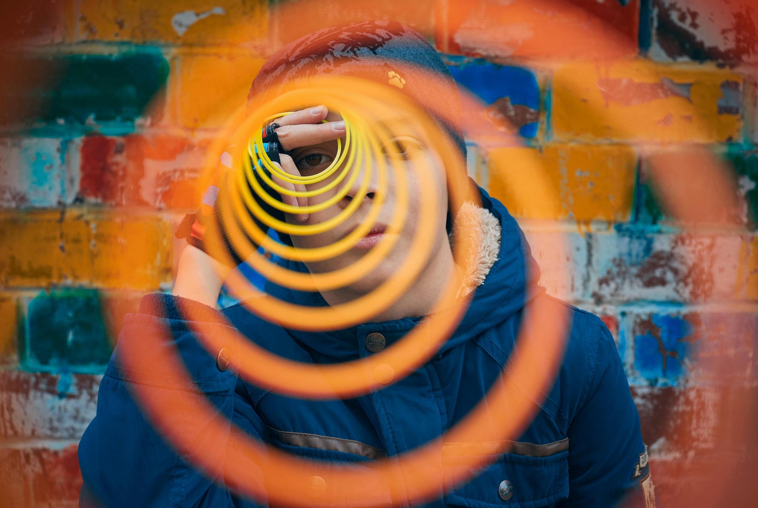 boy standing in front of a brick wall holding outstretched orange slinky up to his eye