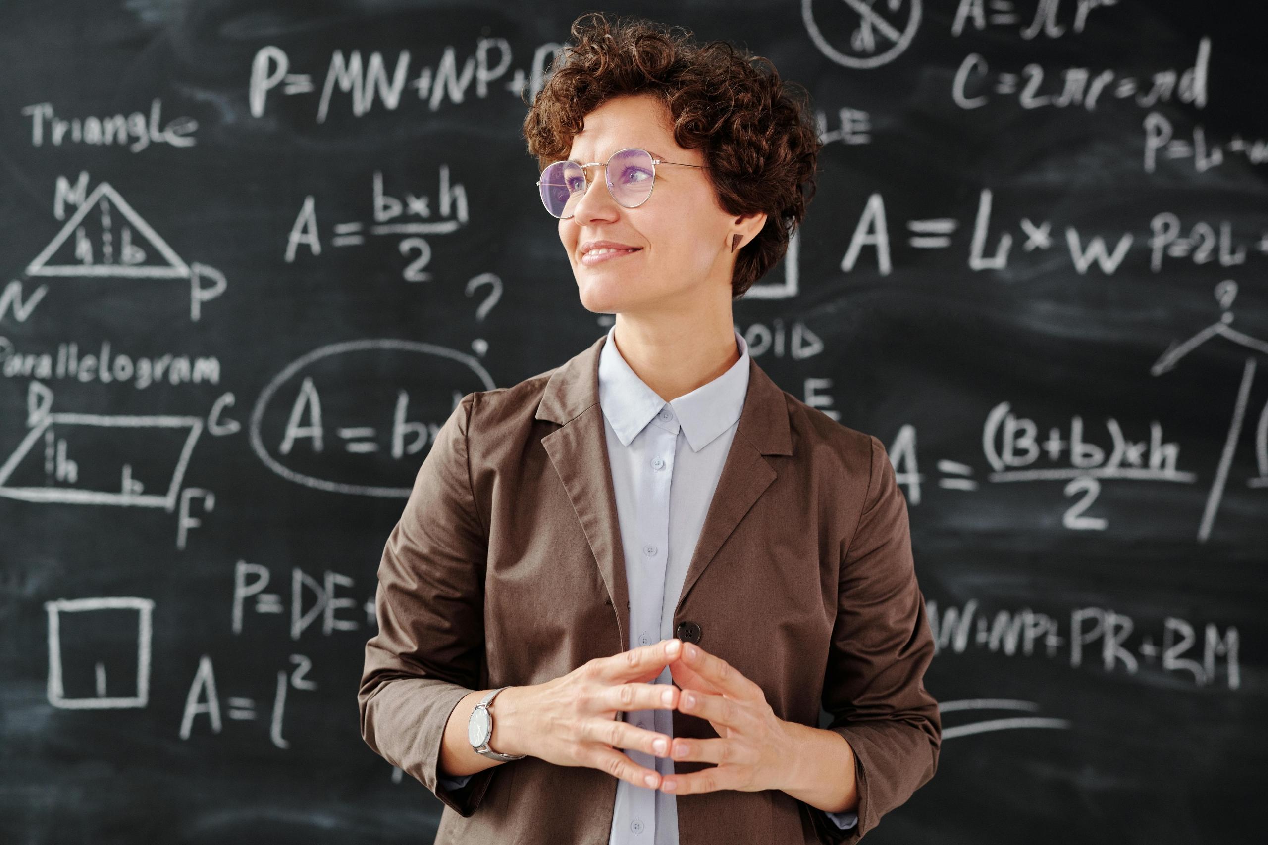 teacher standing in classroom in front of blackboard with maths equations written on it