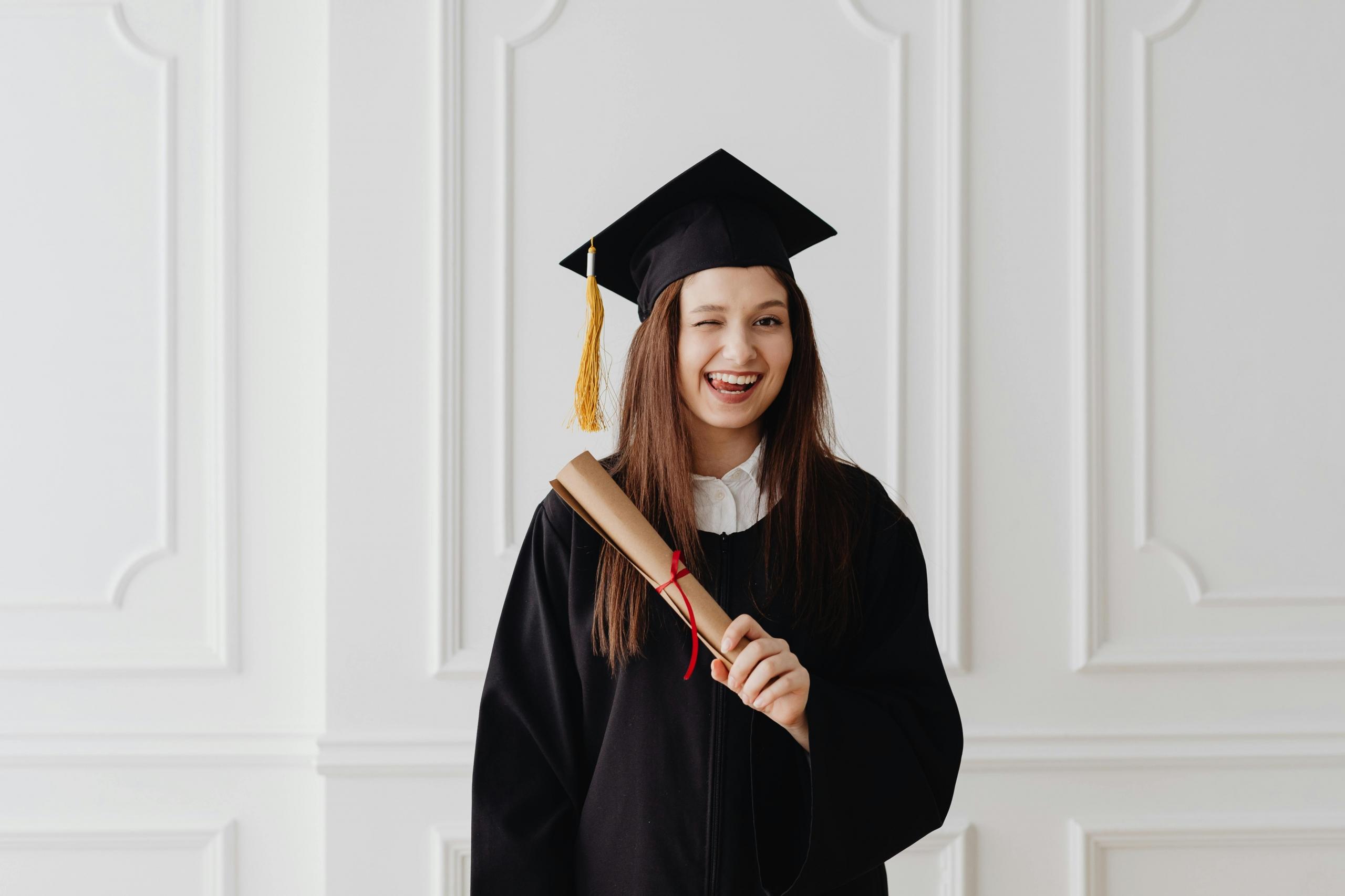 student wearing graduation gown and mortar board holding a diploma and winking