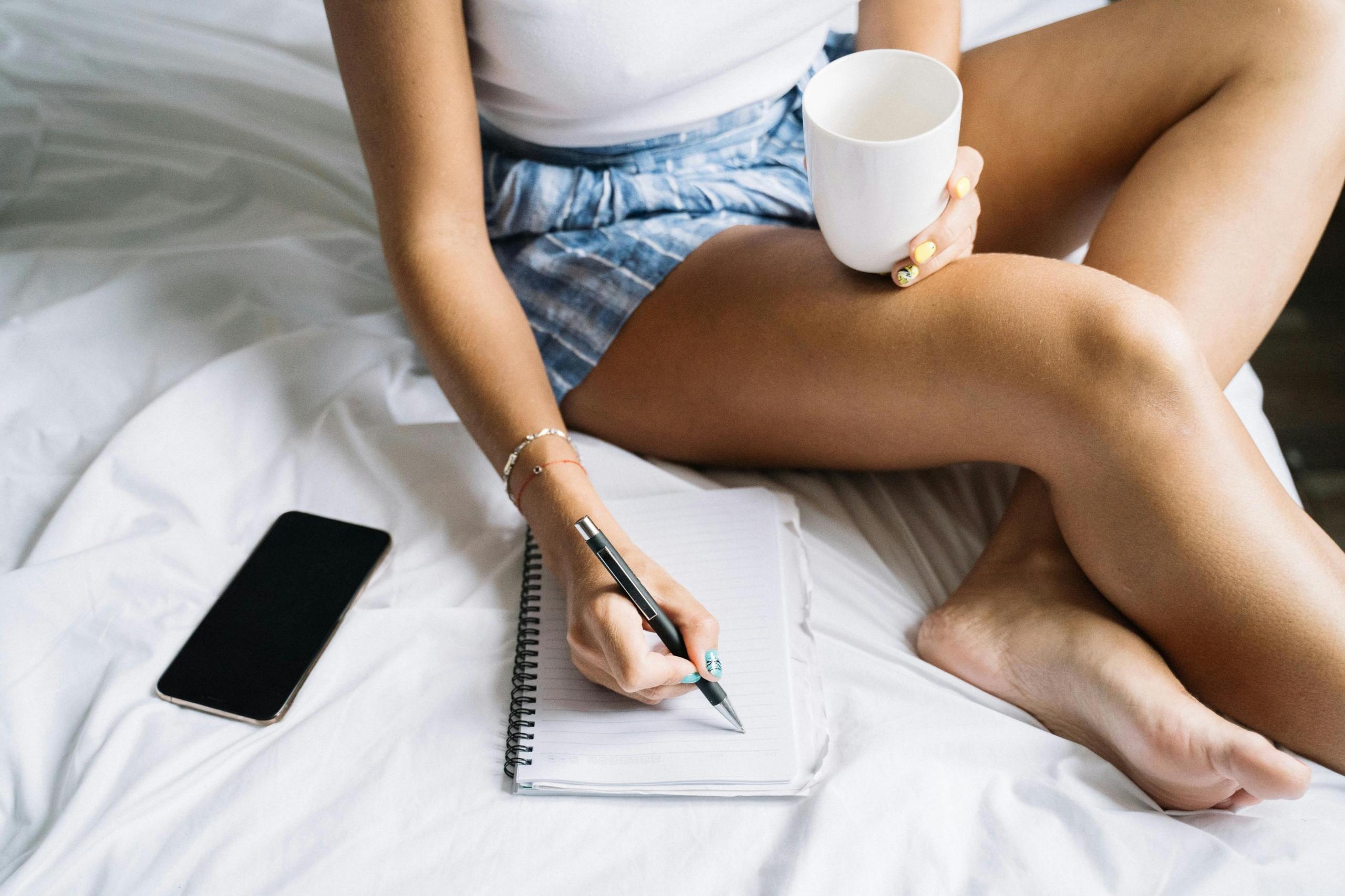 person sitting on bed holding cup of coffee and writing