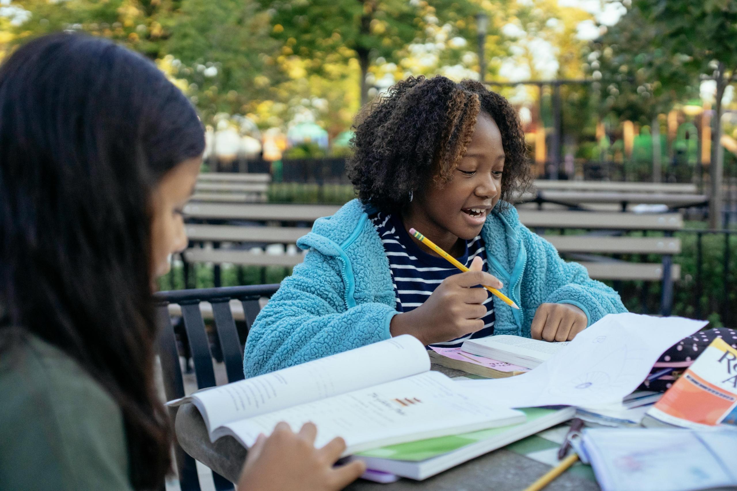 two schoolgirls sitting around outside table writing in books and reading English textbooks