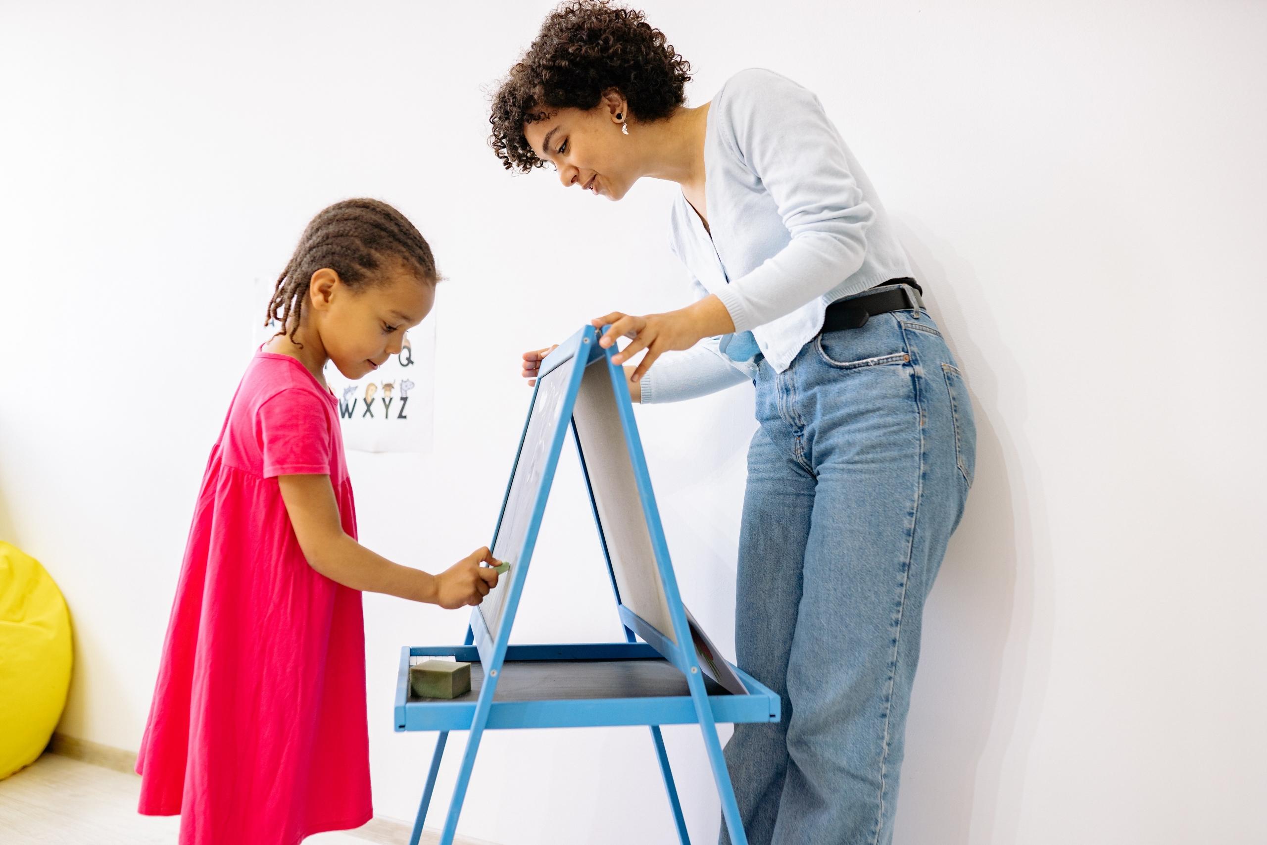 a pre-school teacher and a young girl working on a small chalkboard