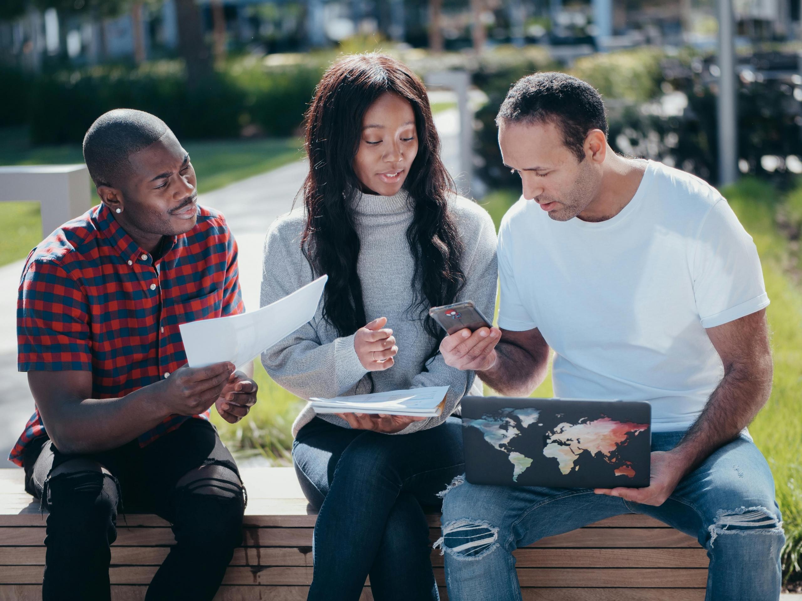 three people sitting side by side on outdoor bench looking at phone, laptop and paperwork