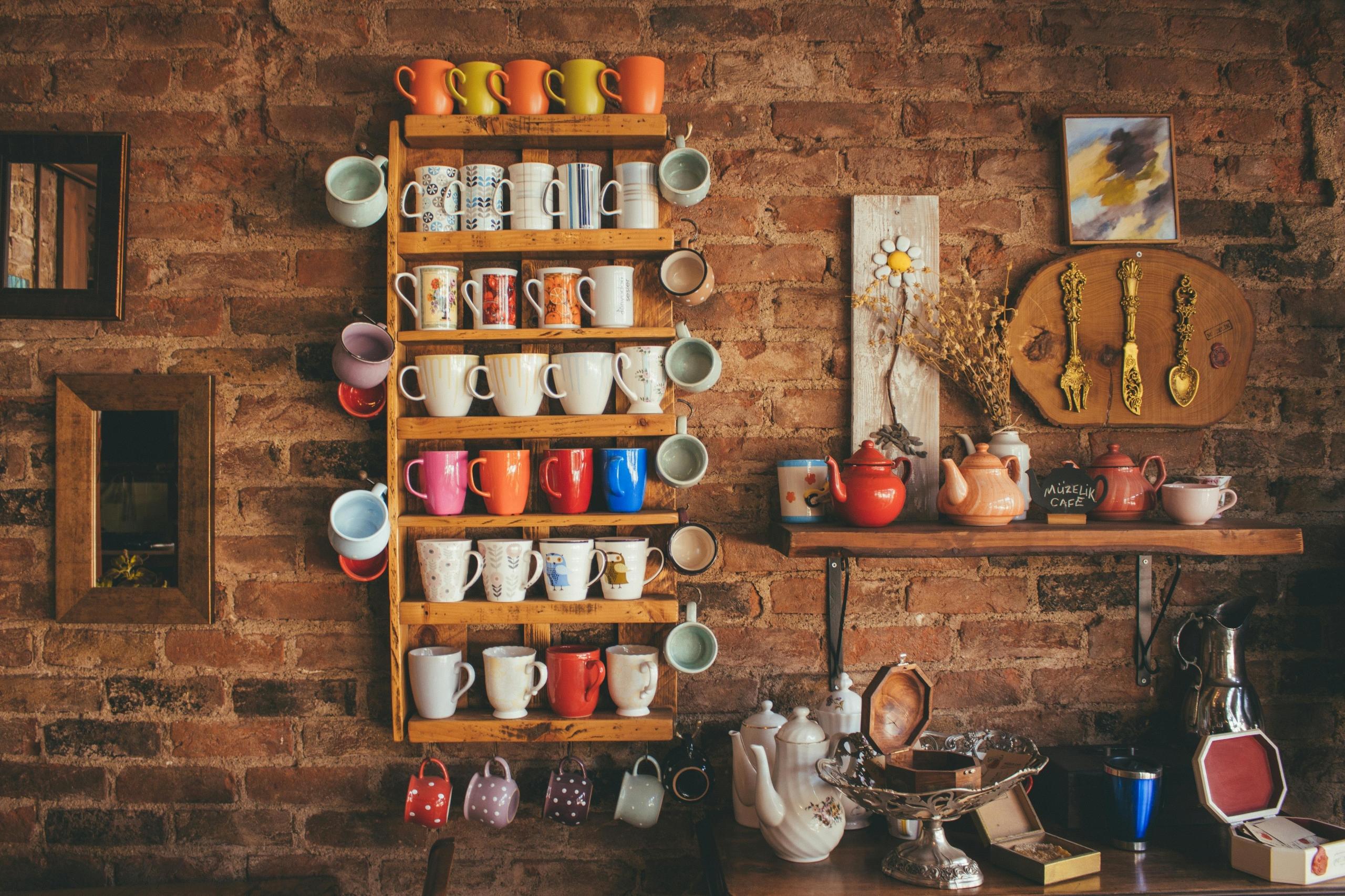 display of colourful mugs on wooden shelf against brick wall near collection of teapots