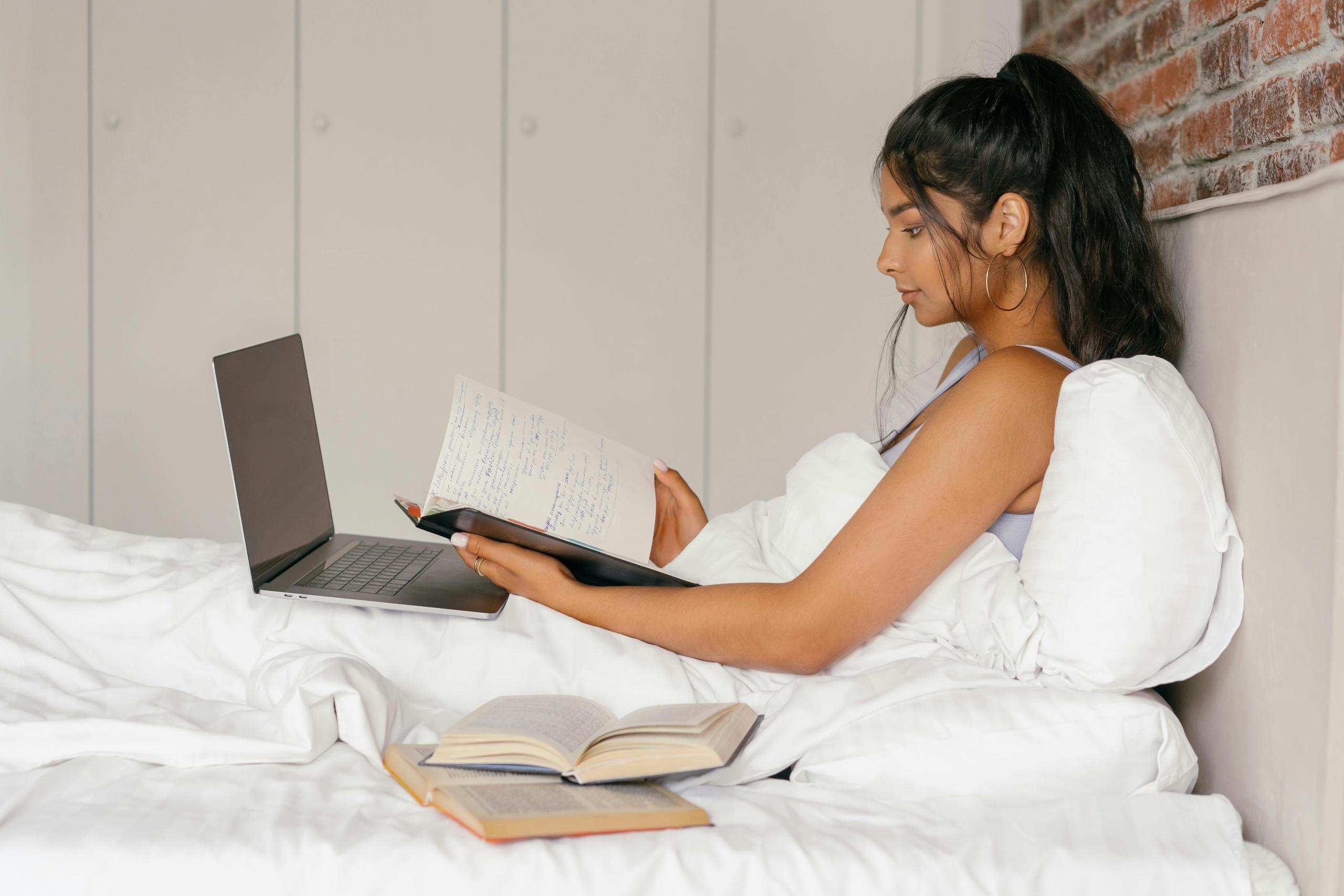 student sitting under duvet cover studying with books and a laptop