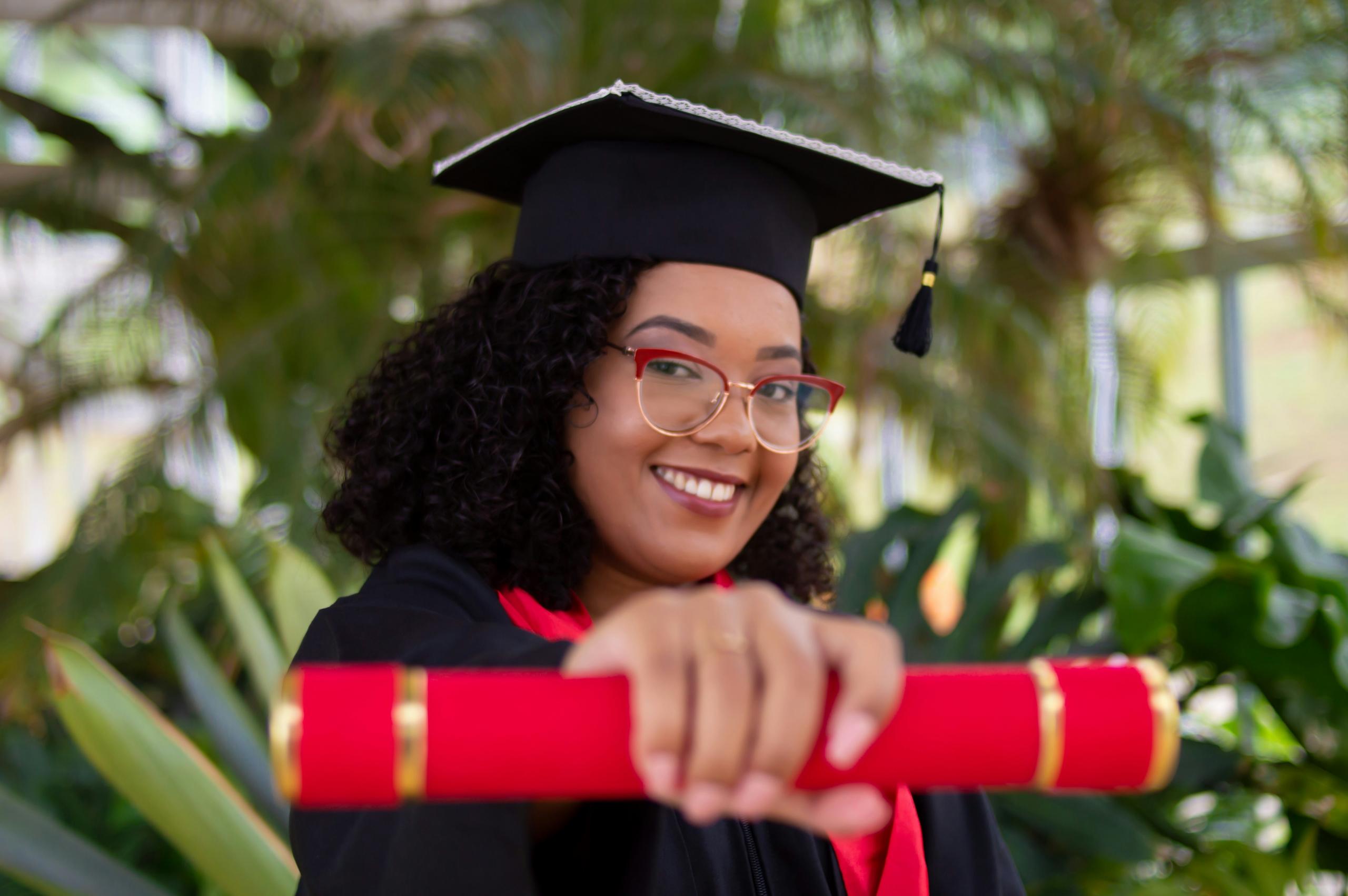 woman in graduation gown and mortar board holding diploma covered with red scroll to camera