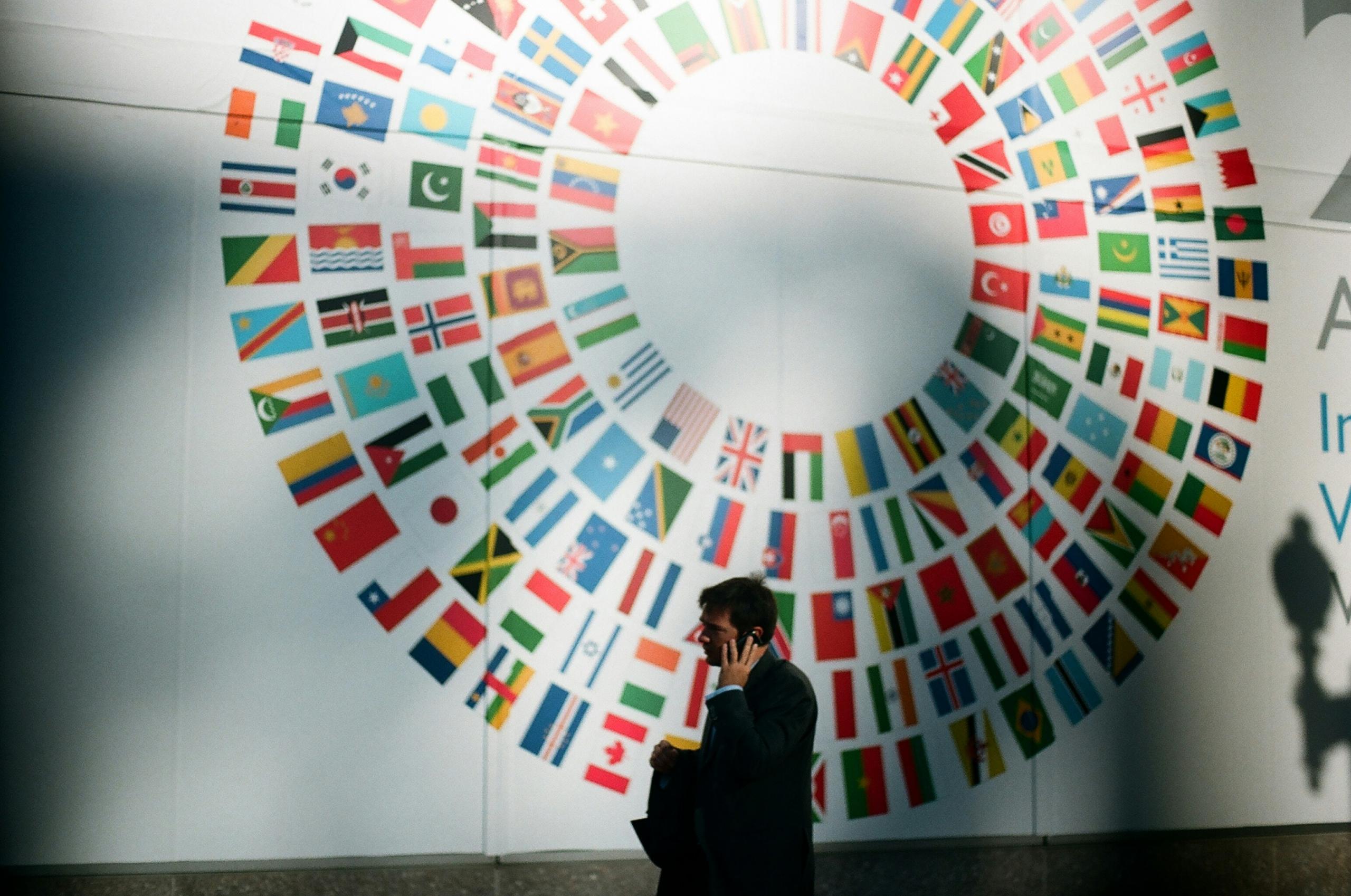 person talking on smartphone, walking past the World Bank Building with country flags depicted on wall