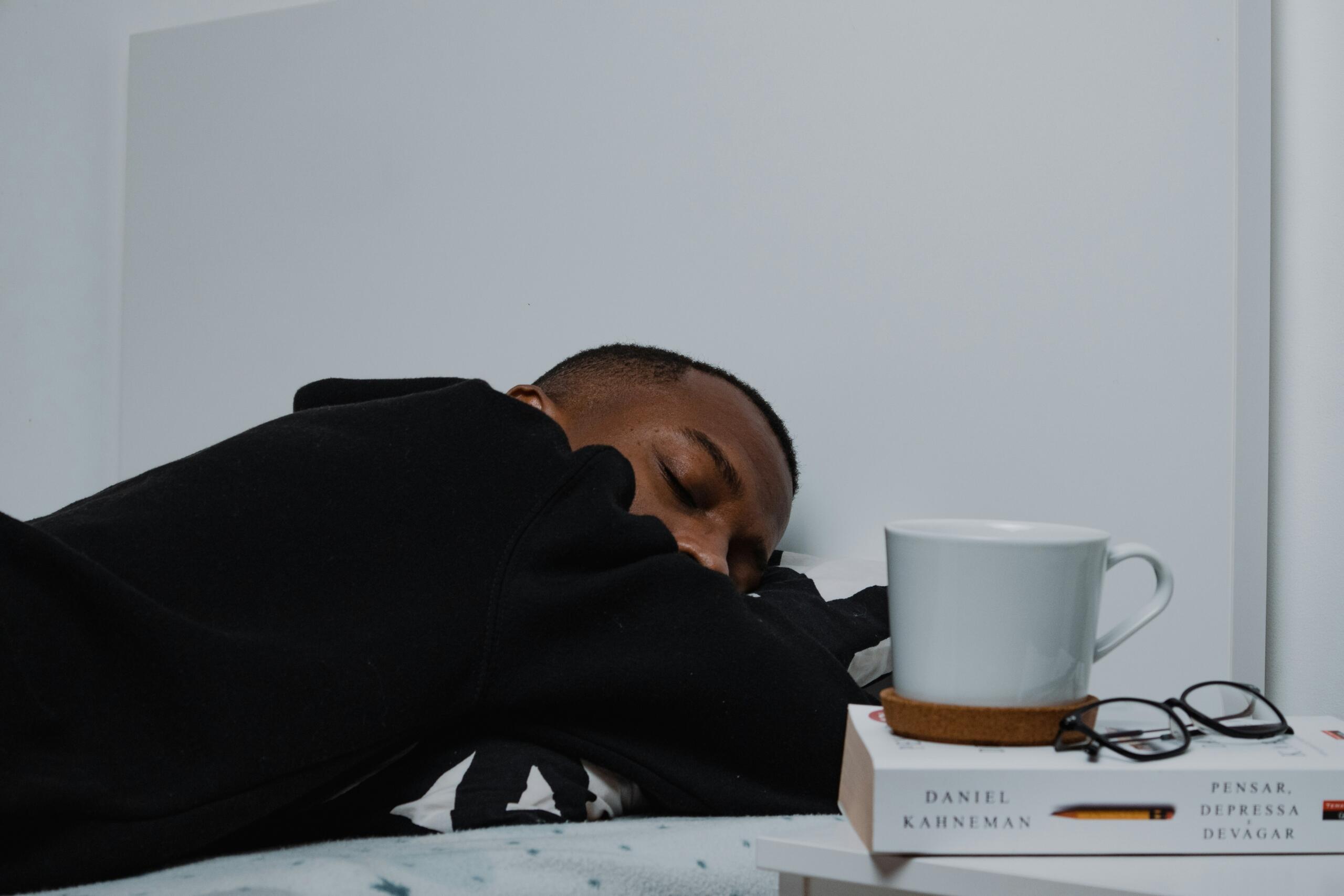 student sleeping on top of bed covers with empty mug and book placed on nightstand