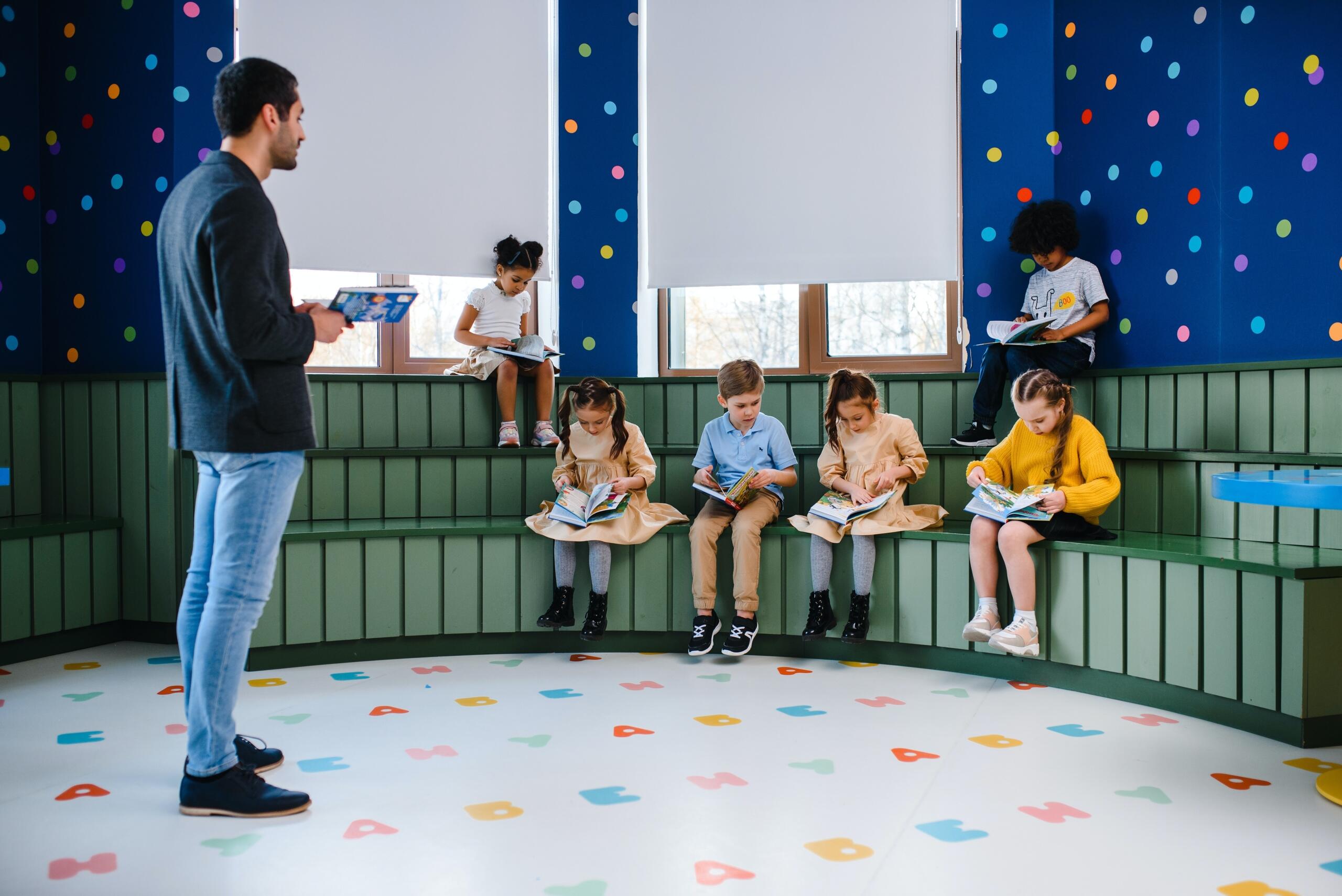 girls and boys sitting on raised benches reading with teacher standing in centre of room