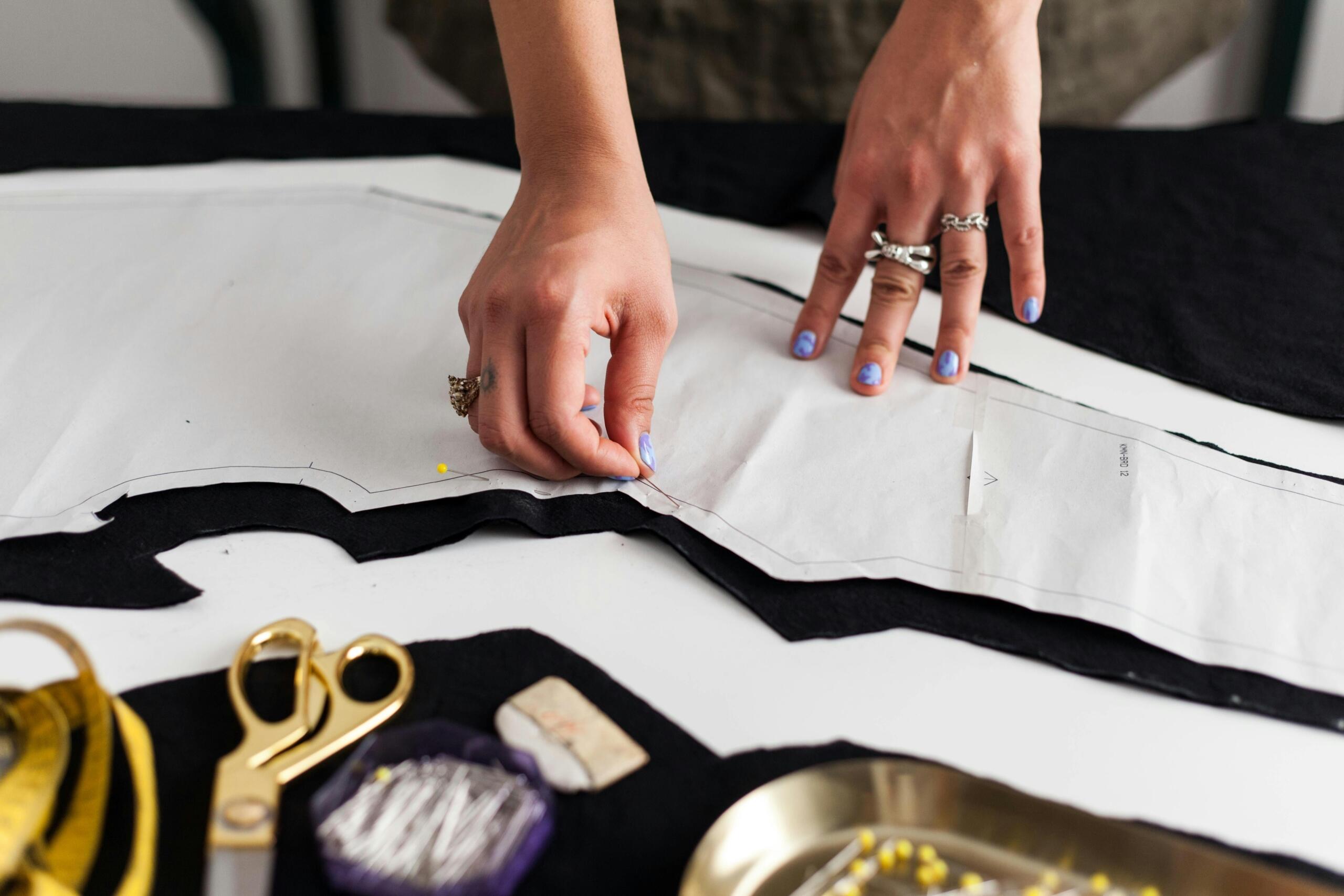 seamstress using ball-point pins to pin a pattern to fabric with dressmaker's scissors on table
