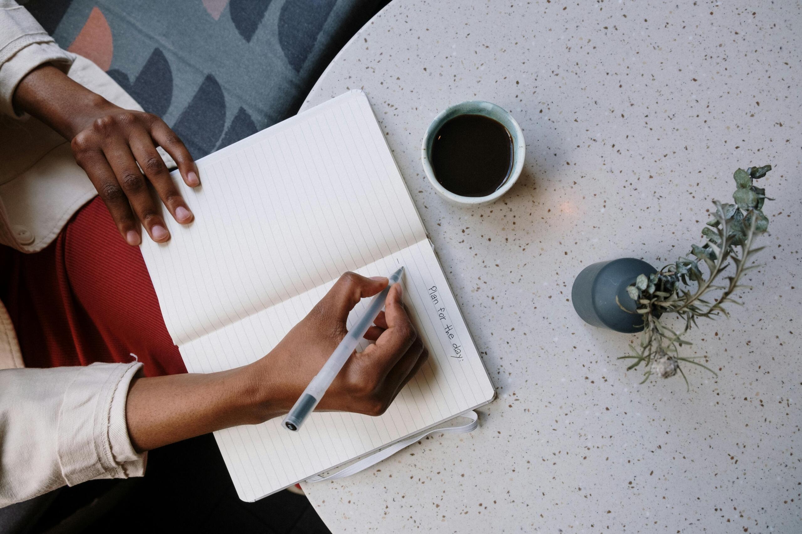 person seated at table drinking coffee and holding black pen to white lined notebook paper